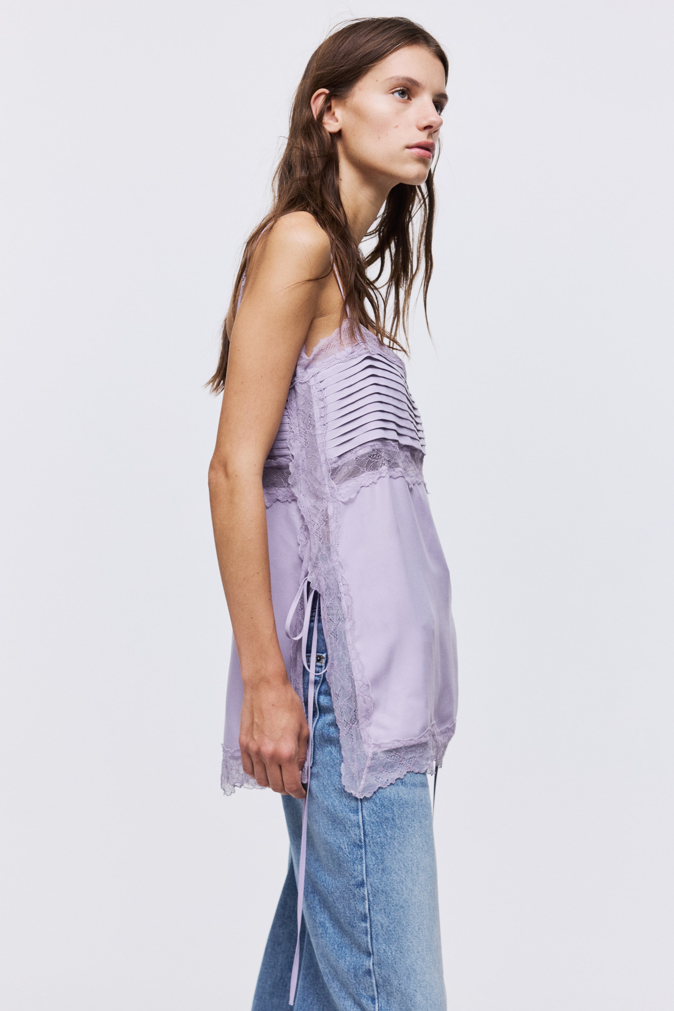 View larger image: A long light purple top, made from a viscose weave, is featuring lace edges, insets, a pleated bust section, and adjustable spaghetti shoulder straps. A high side slit with narrow ties is visible on the side as the model is wearing the top with blue jeans.