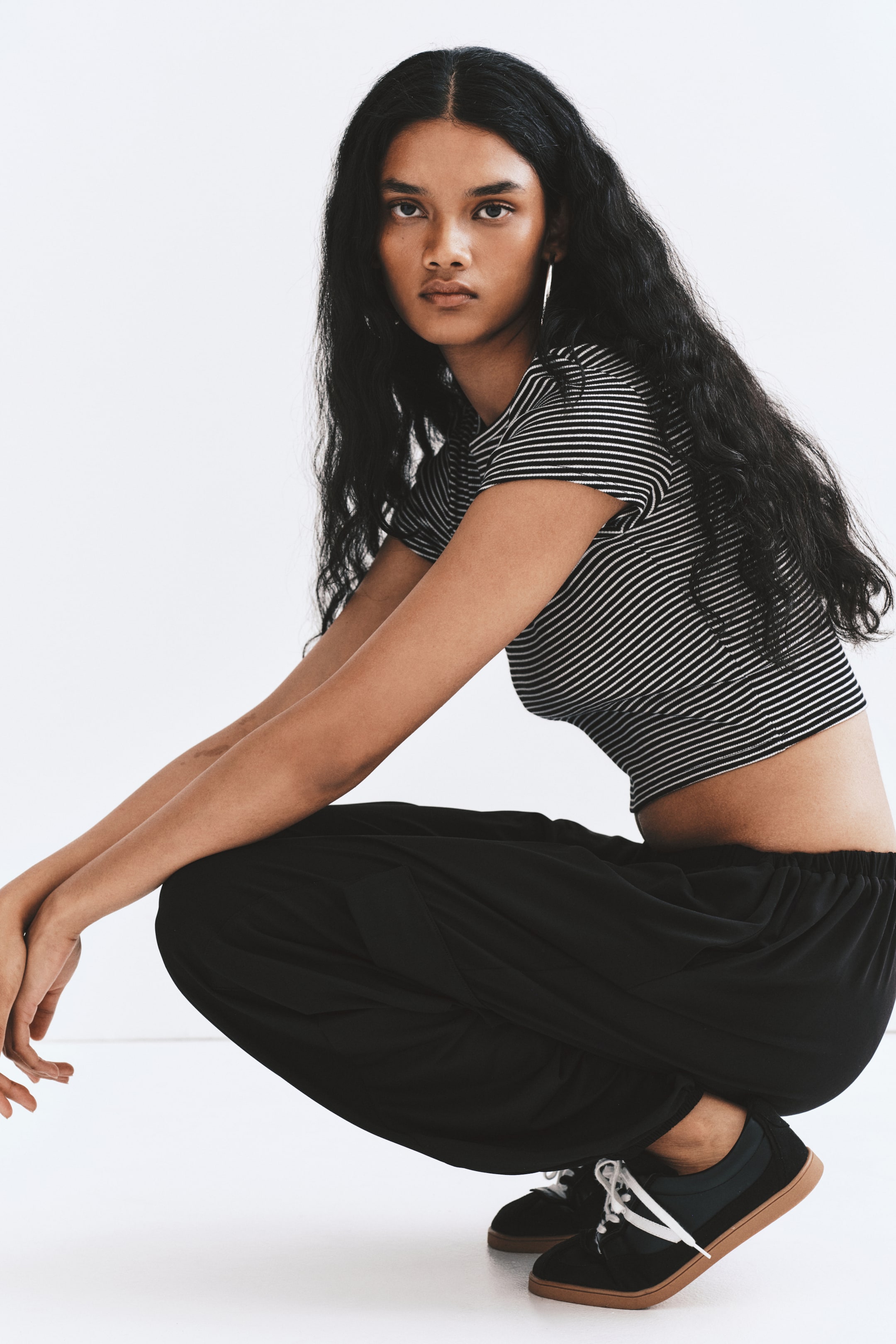 Visualizza l'immagine ingrandita: Pantaloni balloon neri con vita bassa elasticizzata e vestibilità ampia e fluida, indossati dal modello accovacciato. I pantaloni sono abbinati a un crop top a righe bianche e nere e a sneakers nere.