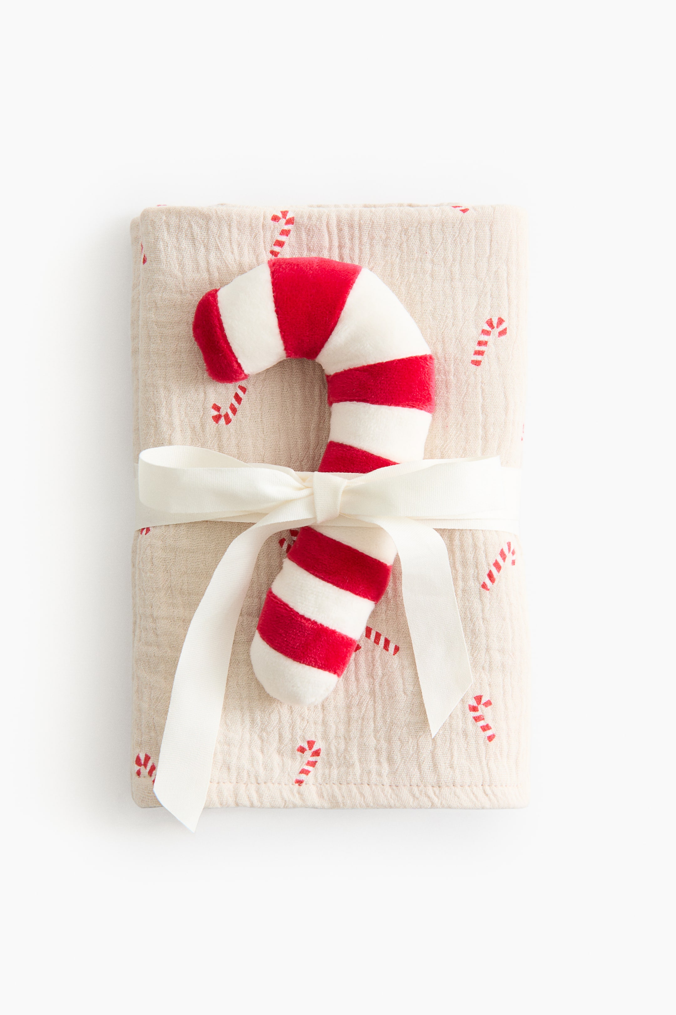 View larger image: Gift set featuring a candy cane-shaped rattle and a folded blanket. The rattle is made of velour and features red and white stripes. The blanket is made of soft cotton muslin and printed with small red candy canes on a light background, and both items are tied with an off-white ribbon.