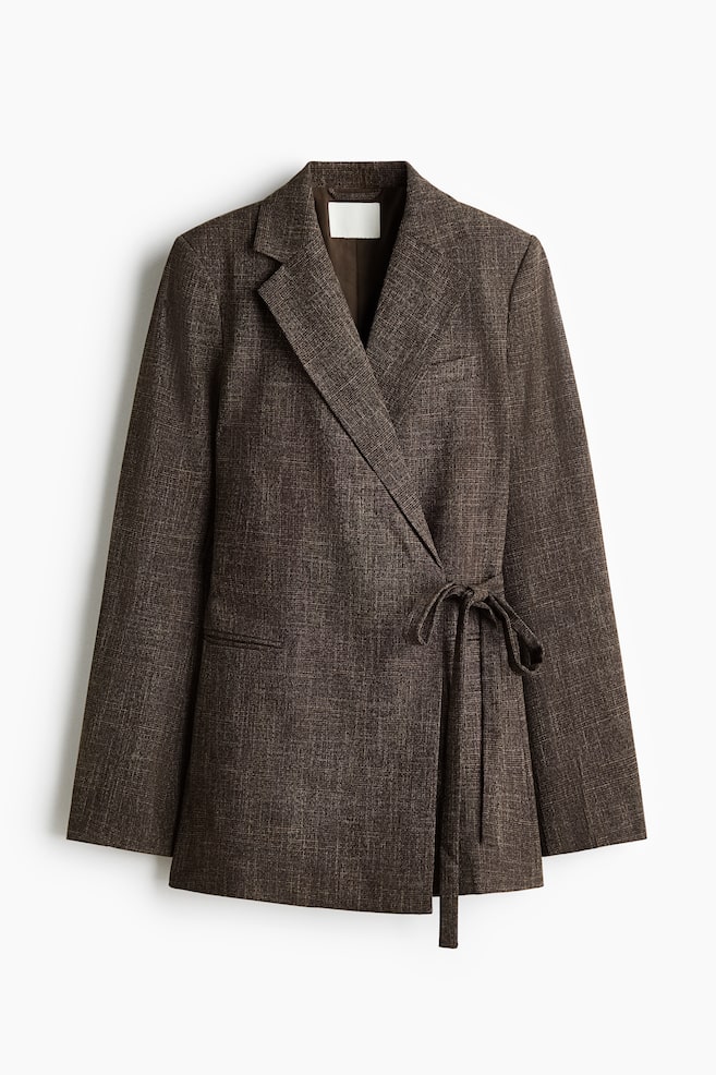 Ein braun strukturiertes Wickelblazer hängt frontal ausgerichtet und zeigt ein dezentes Kreuzschraffurmuster sowie steigende Revers. Der Blazer verfügt über einen Wickelverschluss, der mit einem Bindegürtel aus dem gleichen Stoff in der Taille fixiert ist, sowie lange, gerade Ãrmel. - 1