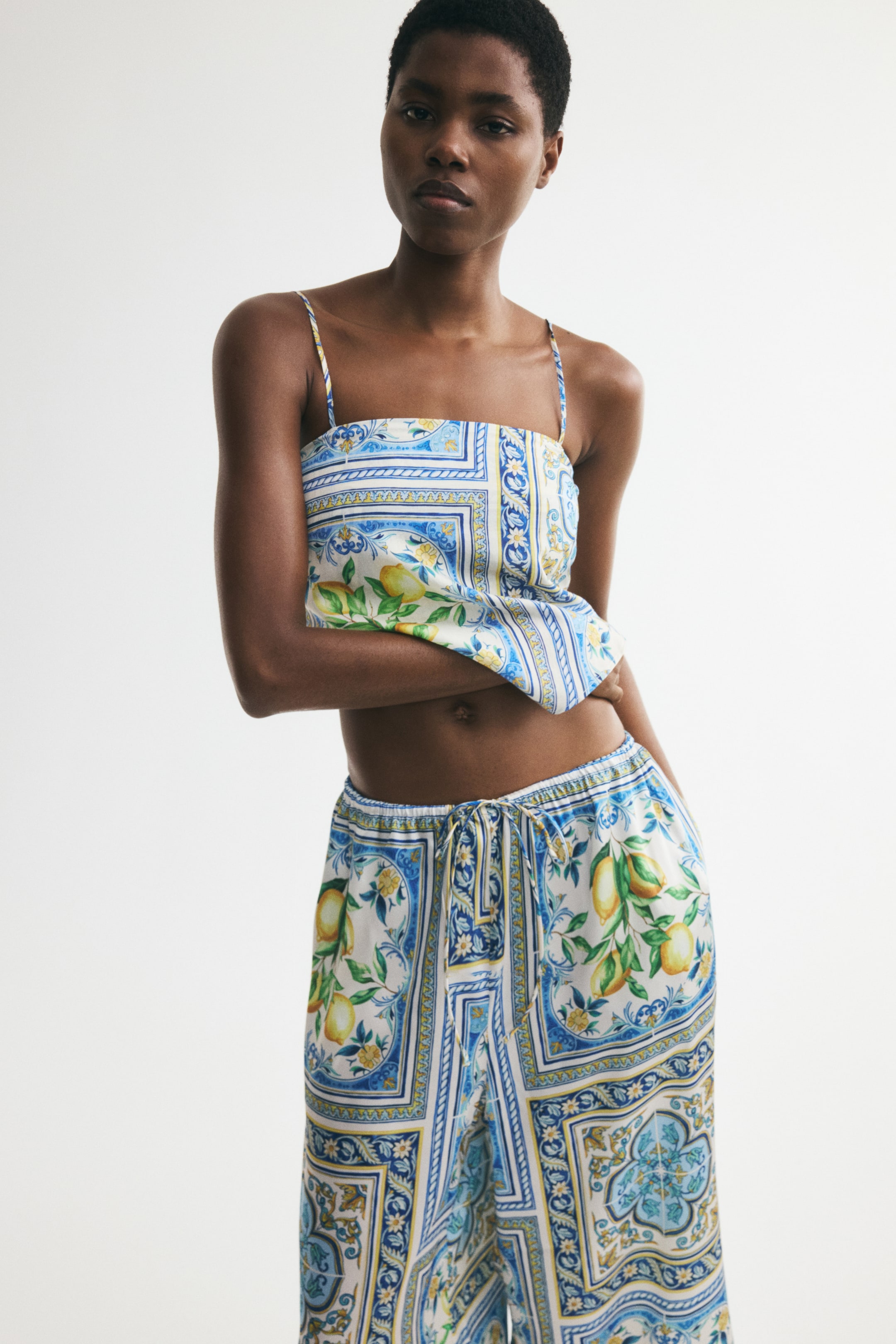 Agrandir l'image: Un haut court en tissu mélangé de viscose imprimé, avec un décolleté droit, des bretelles fines et un ourlet en forme de V à l'avant, est porté par le mannequin. Le haut présente un imprimé bleu, blanc et jaune avec des motifs de citrons et des motifs en carreaux, et est assorti à un pantalon imprimé coordonné.