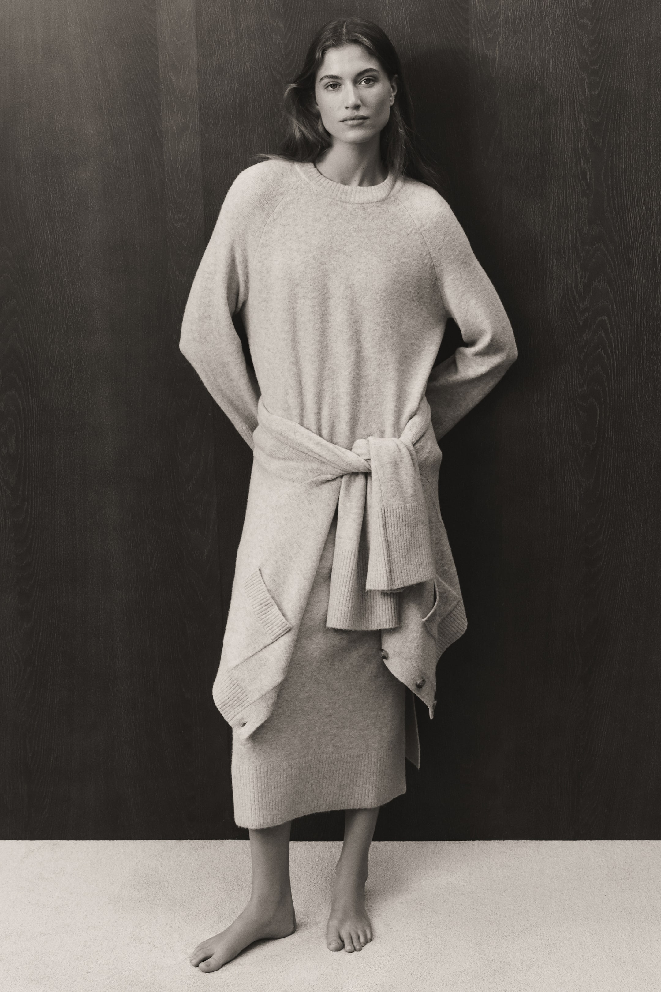 View larger image: A knit maxi dress is shown from the front, featuring a round, rib-trimmed neckline, long raglan sleeves, and a straight ribbed hem. A matching knit cardigan is tied around the waist, highlighting the dress’s soft texture and relaxed fit.