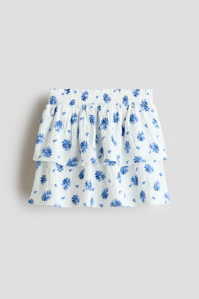 La falda muestra una tela blanca arrugada con un estampado floral azul. Presenta una pretina fruncida y dos niveles con volantes, creando una silueta voluminosa en el frente. - 1