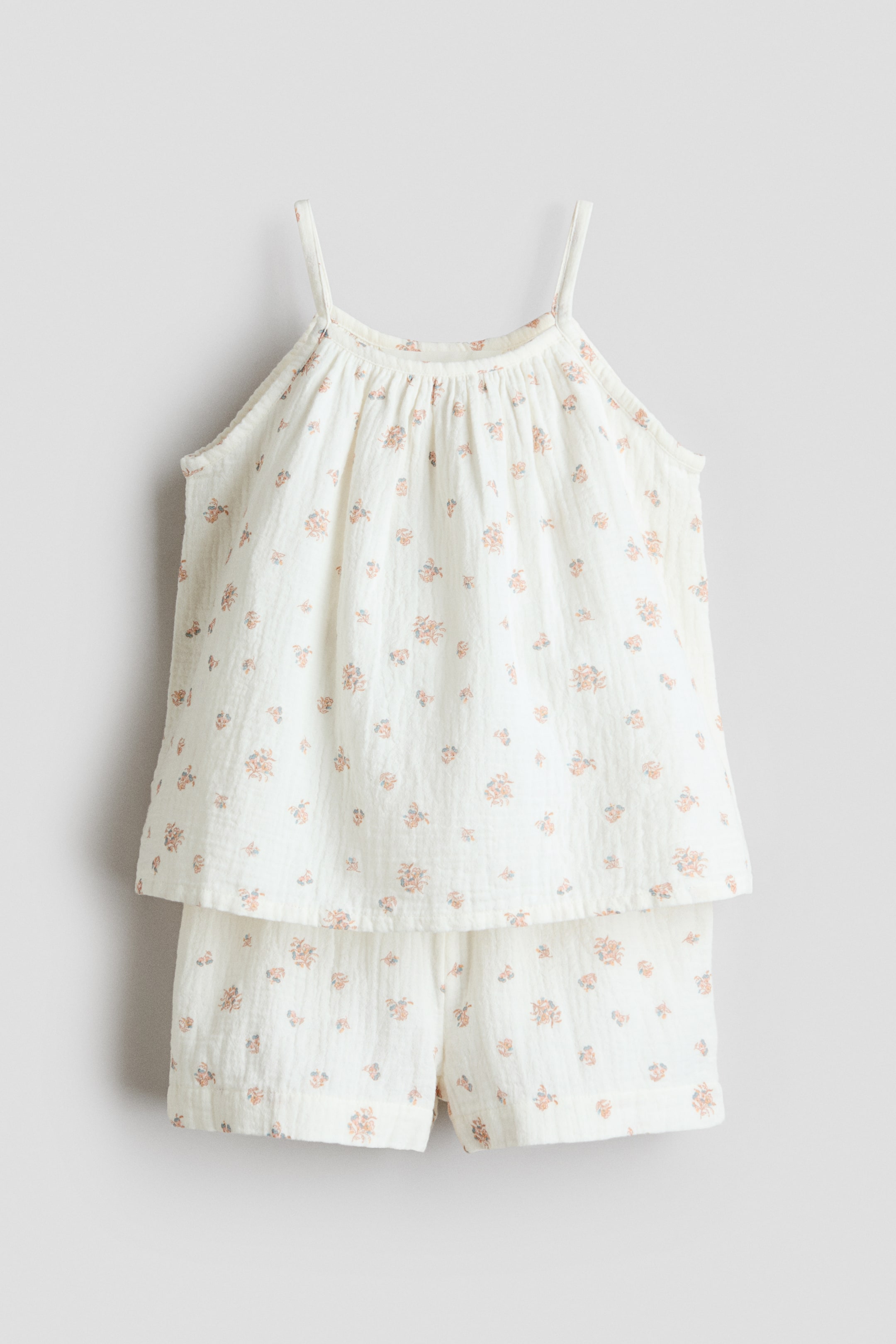 Grotere afbeelding bekijken: Een H&M Adorables set, bestaande uit een top met bandjes en shorts in off-white mousseline, wordt van de voorkant getoond. De top heeft een uitlopende snit met een gerimpelde ronde halslijn en smalle schouderbandjes, terwijl de bijpassende shorts een elastische taille hebben. Beide stukken zijn versierd met een patroon van kleine perzik- en blauwe bloemen.