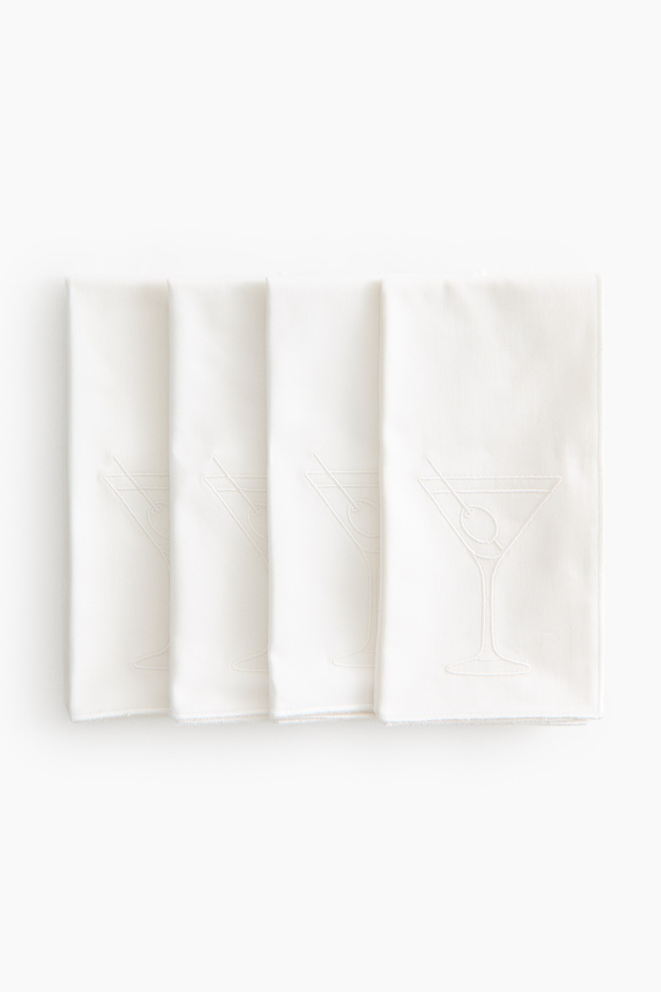 View larger image: Four off-white cotton sateen napkins are lying flat in a row, overlapping slightly to show their front designs. Each vertically folded napkin is displaying a satin-stitched martini glass with an olive and matching stitched borders.