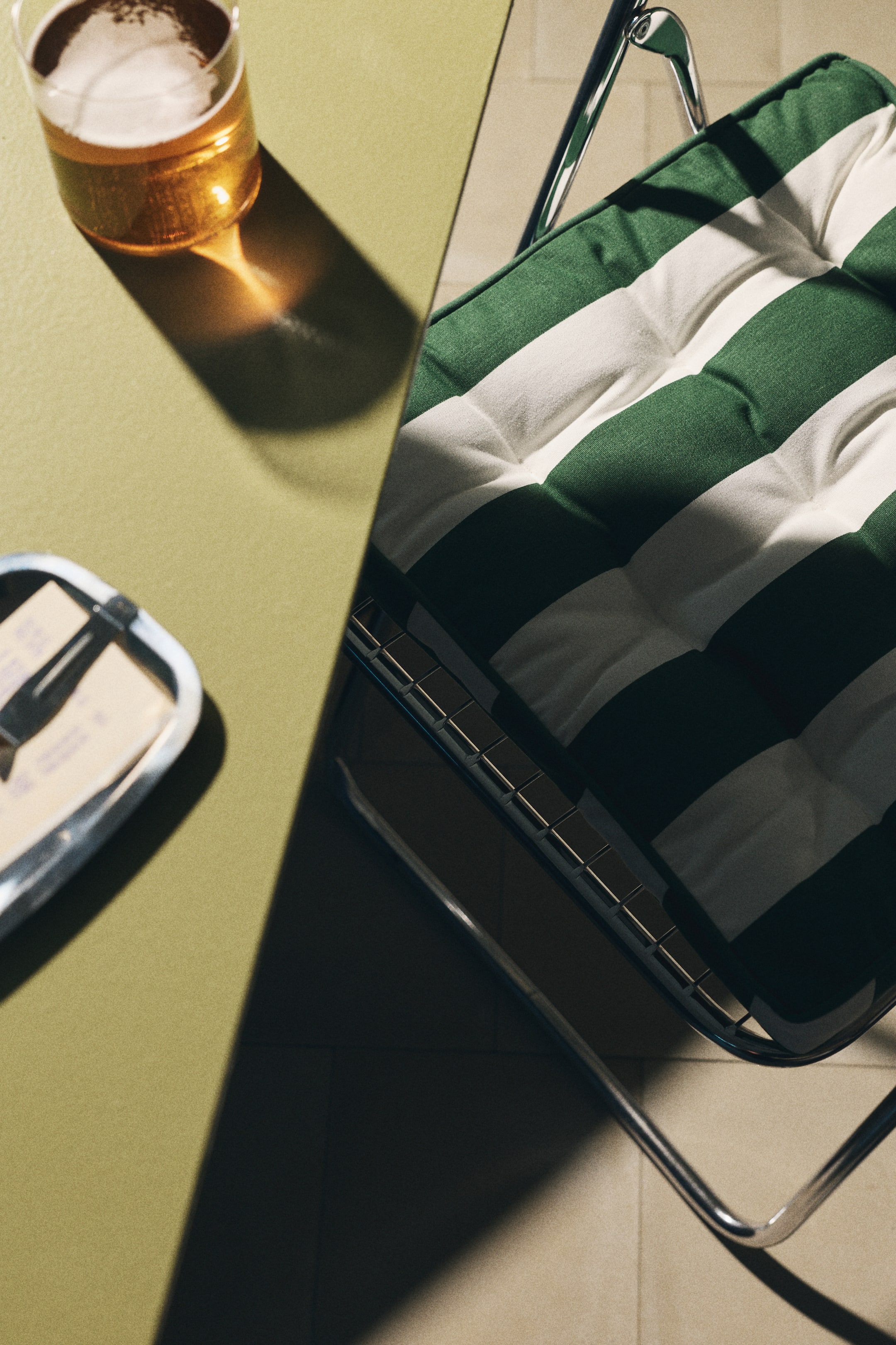 Ampliar la imagen: Una silla con un cojín capitoné, que presenta franjas alternas anchas en verde y blanco. El cojín descansa sobre un marco curvado de metal tubular y muestra profundas hendiduras debido a su capitonado.