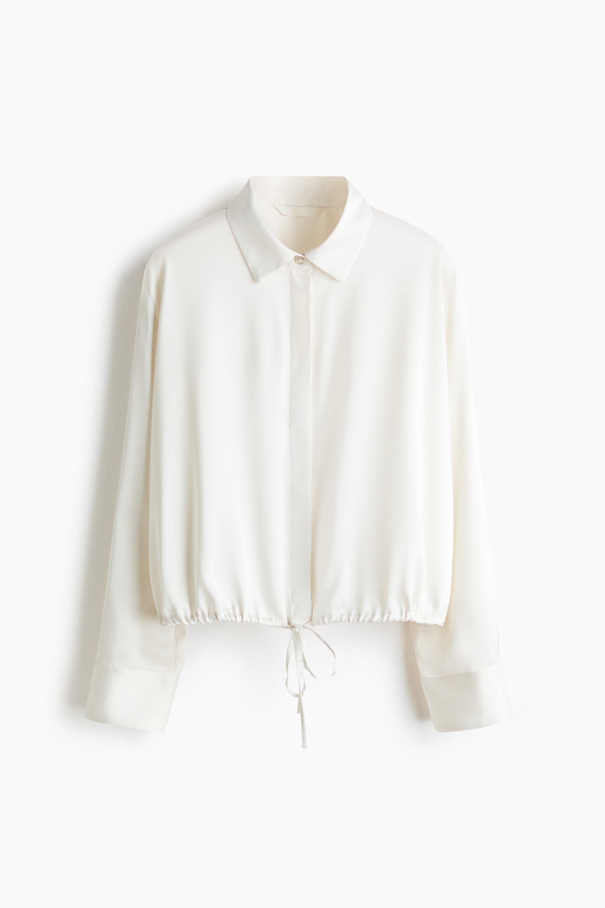 Una modelo lleva una camisa de satén blanco de caÃda suave, con cuello italiano, botones delanteros ocultos y mangas largas con puños abotonados. La camisa se muestra de frente, con un cordón blanco en el bajo atado a la cintura sobre unos shorts de piel negros.
