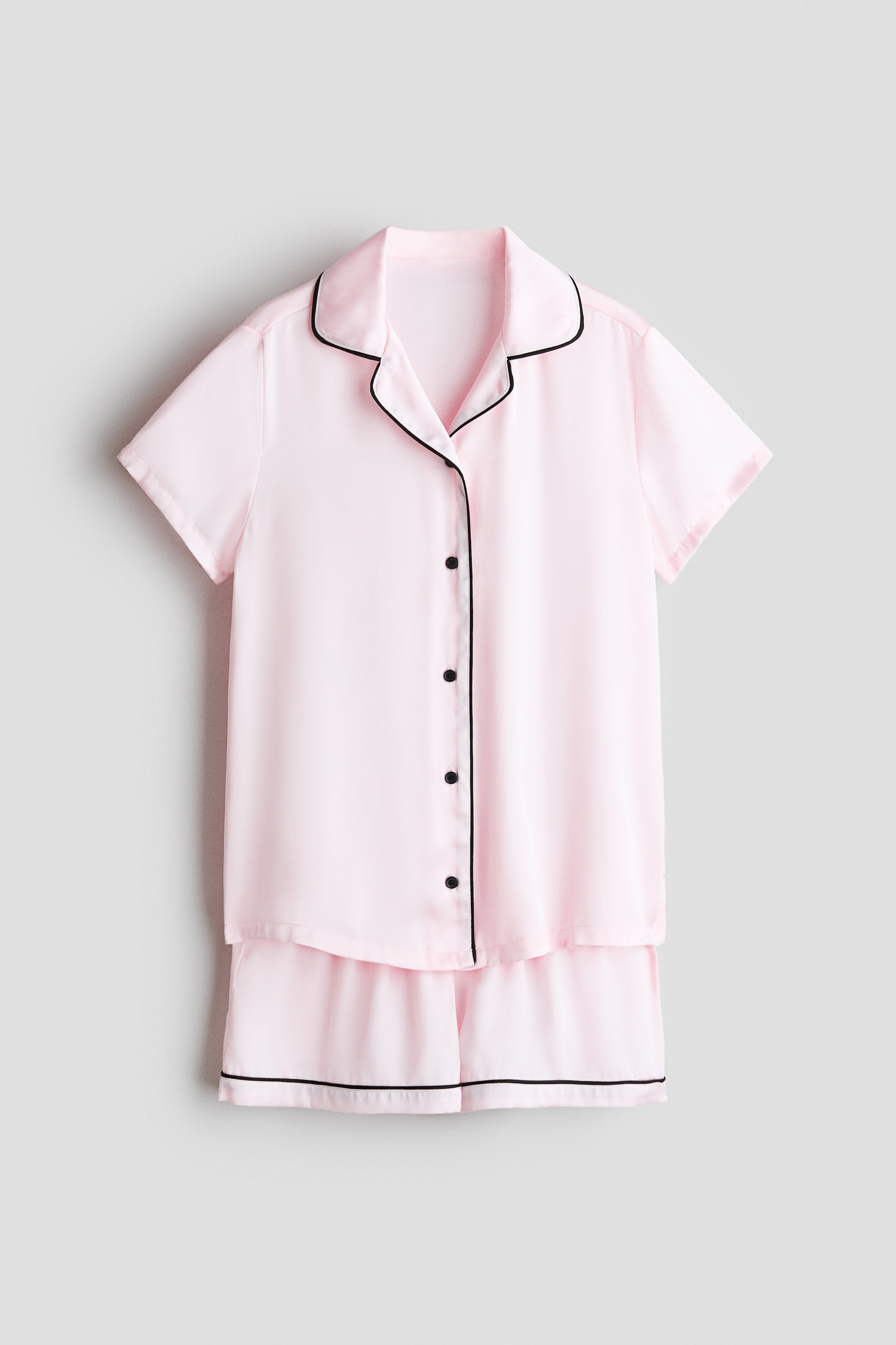View larger image: Pyjamas with a shirt and a pair of shorts in soft pink satin are showcased FRONT. The short-sleeved shirt features a collar, black buttons down the front, and black piping along the collar, placket, and sleeve cuffs, while the matching shorts display black piping along the hem.