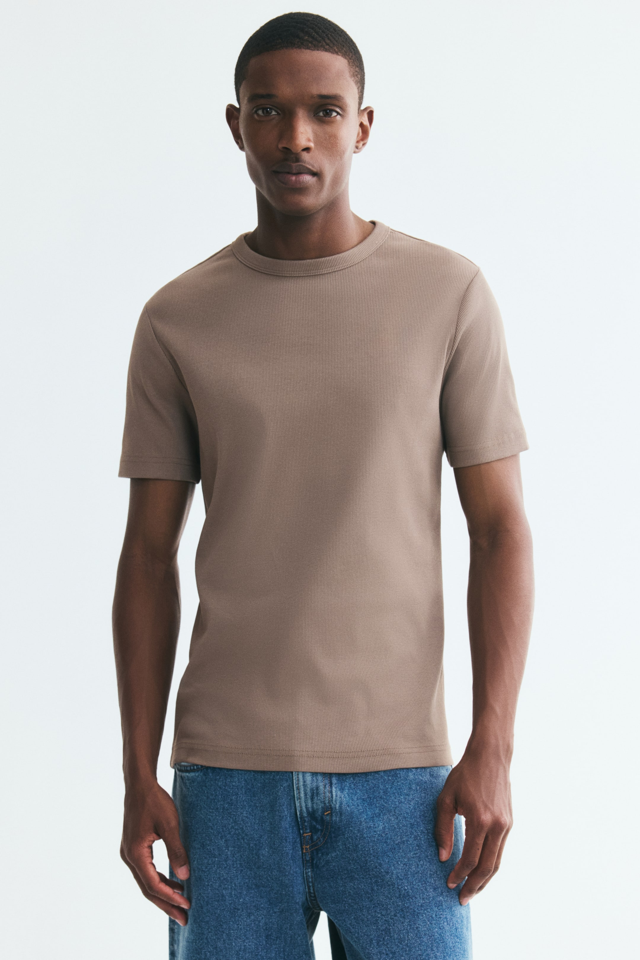 View larger image: The model is wearing a light brown ribbed cotton T-shirt, featuring a slim-fit silhouette, a round neckline, and short sleeves. The T-shirt is paired with blue denim jeans.