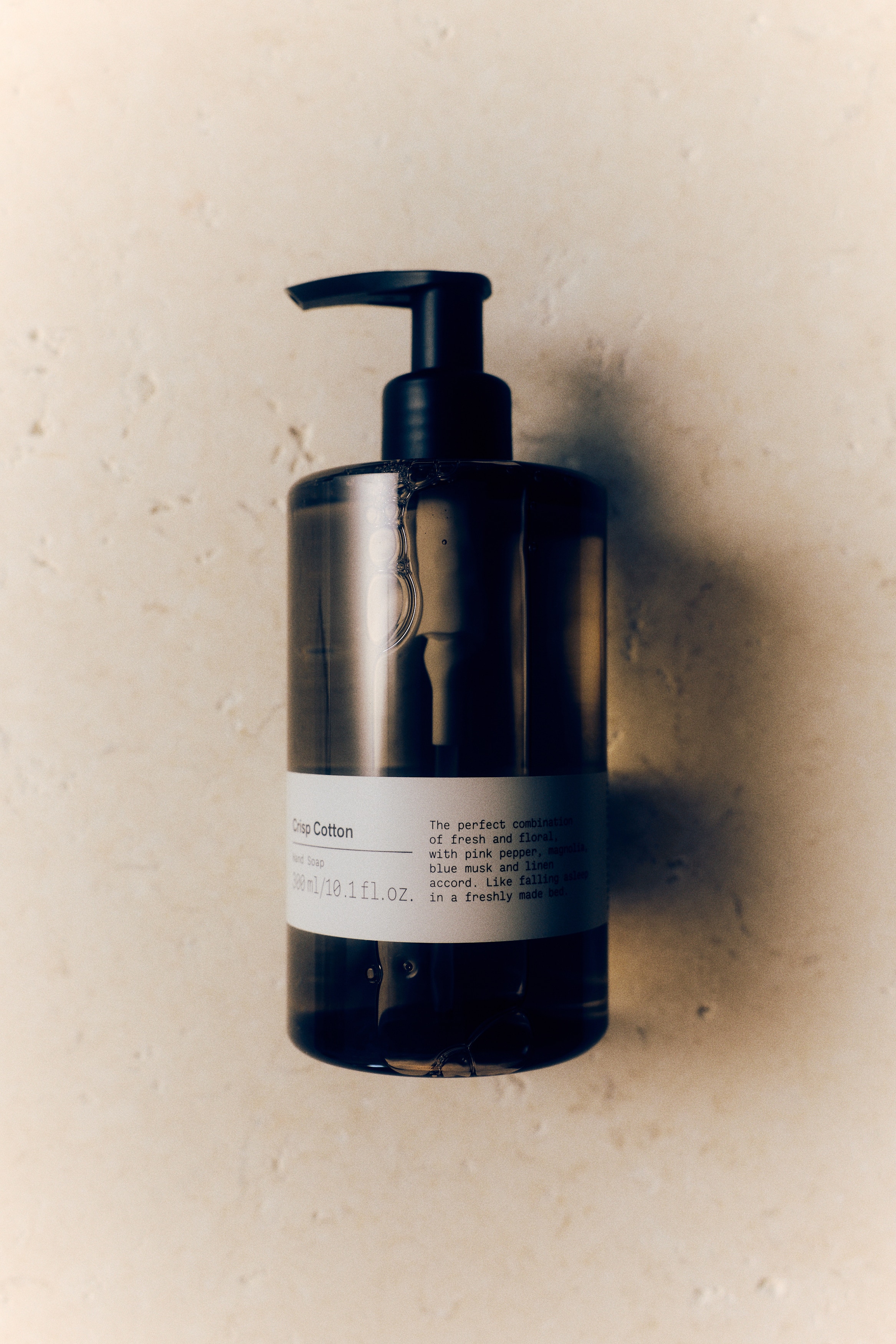 A cylindrical bottle of Crisp Cotton hand soap is standing upright, facing front, featuring a translucent, tinted brown plastic body filled with clear liquid. The bottle is topped with a black pump dispenser and features a white paper label wrapped around the lower half displaying the product name, volume, and description in black text.