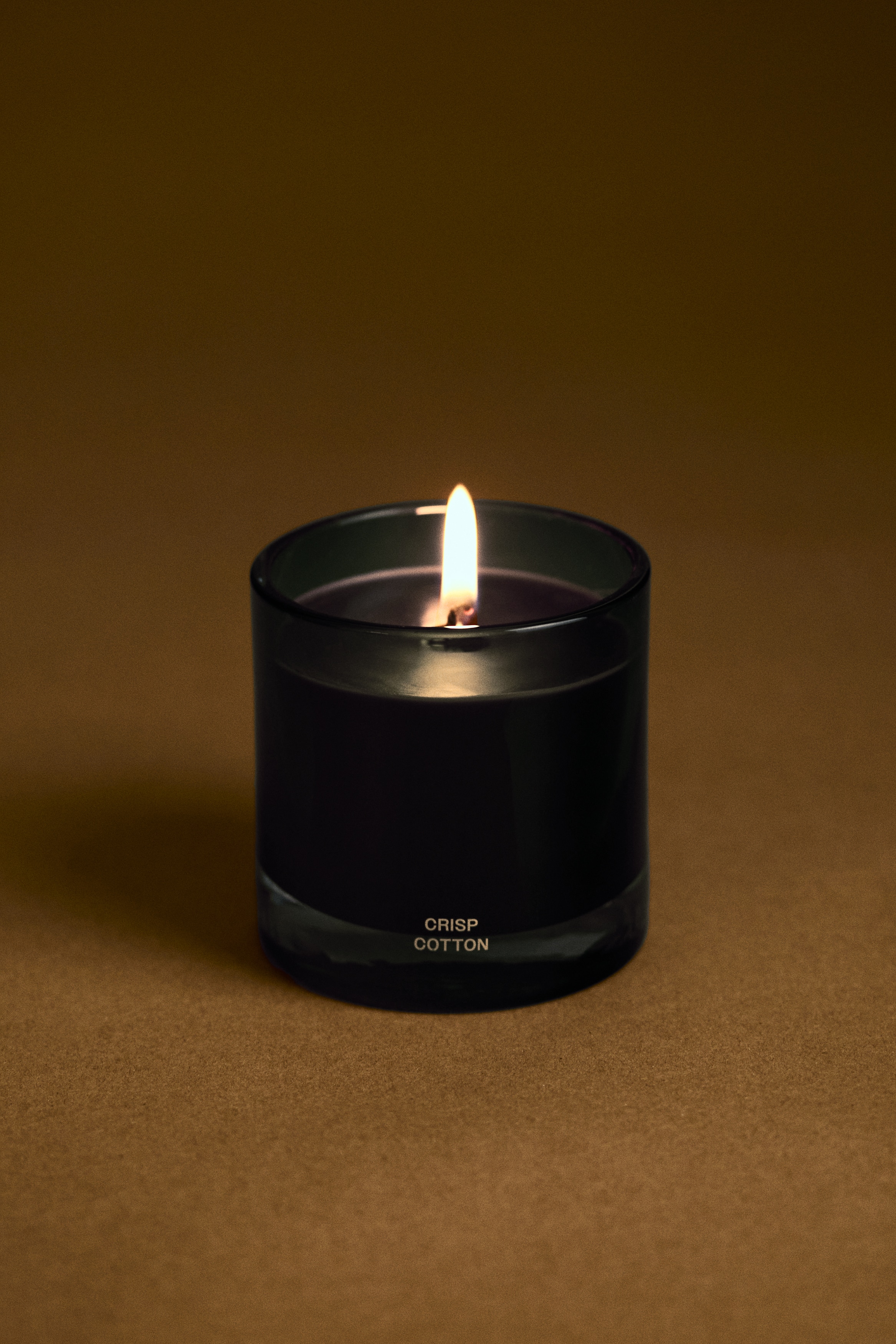 A scented candle stands facing forward in a cylindrical, translucent blue glass container with a thick, clear base. The container holds dark wax with a single central wick and features gold lettering reading âCRISP COTTONâ on the lower front.