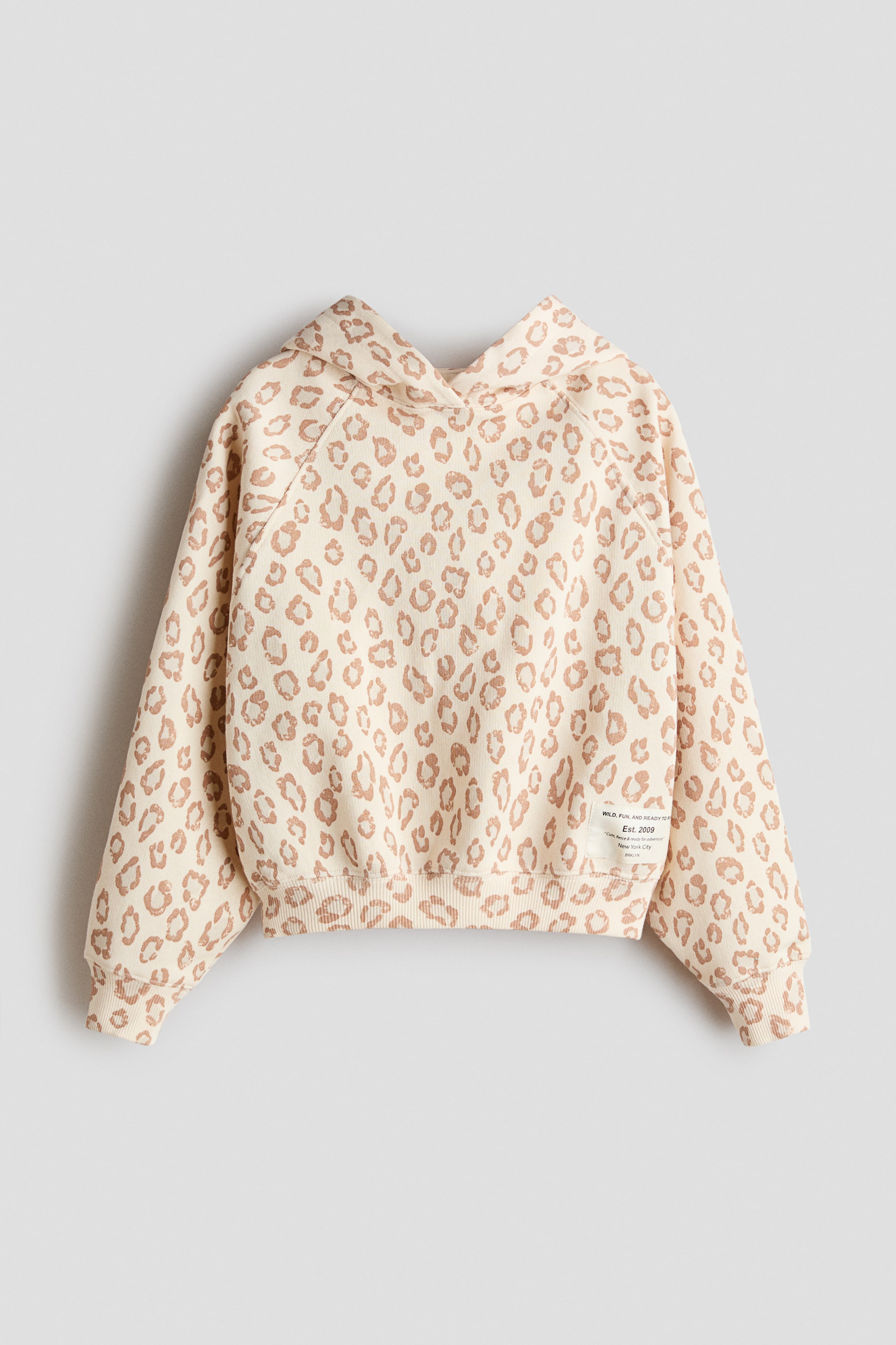 View larger image: A cream hoodie with a light brown leopard print is shown facing forwards. It features raglan sleeves, ribbed cuffs and a ribbed hem. A small rectangular fabric patch is visible on the lower right-hand front.