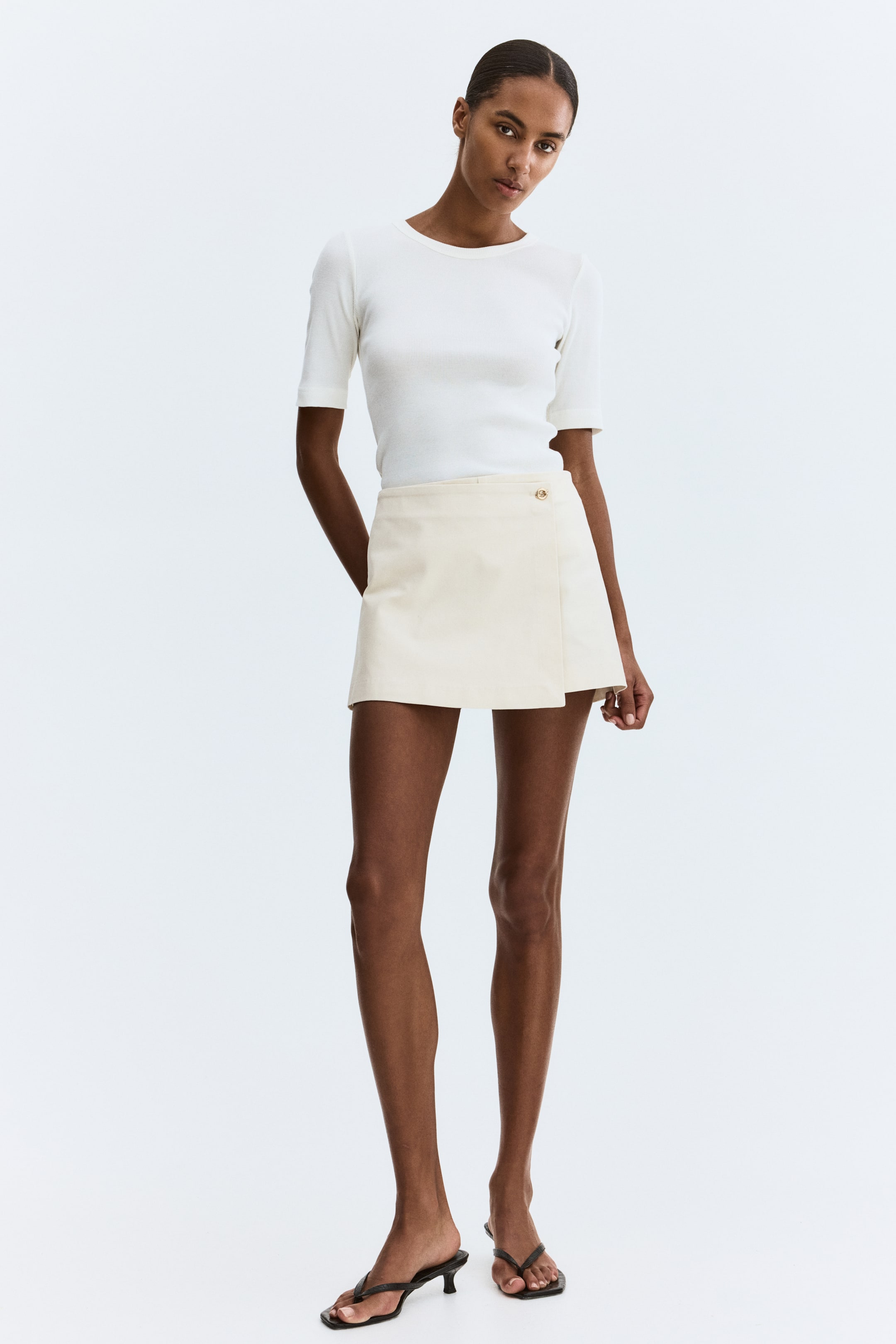 Ampliar la imagen: Un top blanco de manga corta acanalado con cuello redondo y un pequeño emblema oscuro en el lado izquierdo del pecho, con el dobladillo metido en una minifalda envolvente color crema. La falda, vista de frente, presenta un cierre de botón dorado en la cintura y un dobladillo asimétrico.