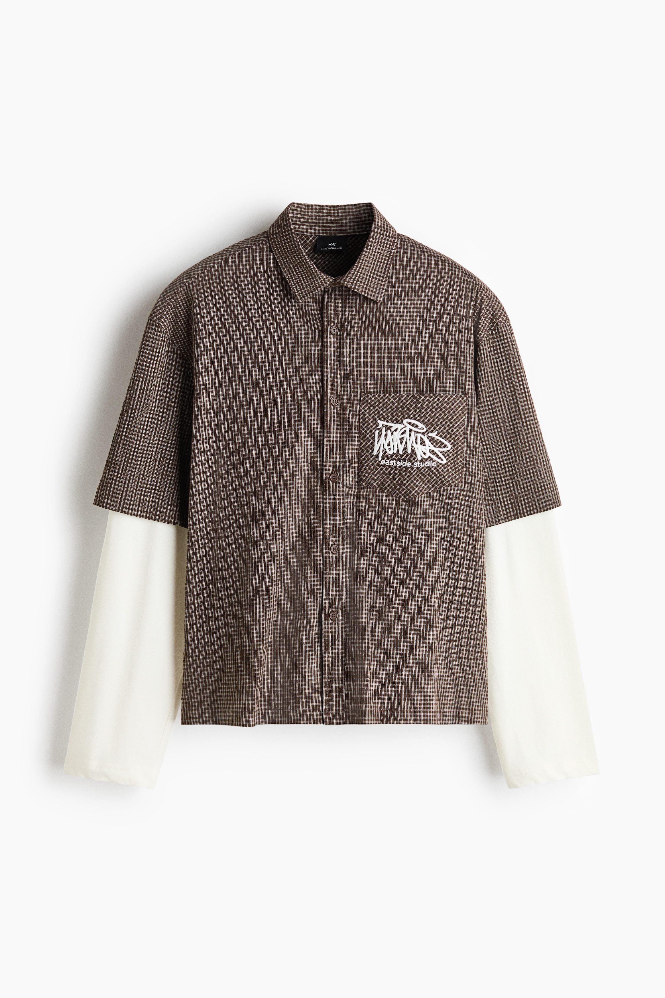 A brown gingham-patterned short-sleeve shirt featuring a white embroidered graphic on the chest pocket is being worn by the model over integrated white long sleeves. The relaxed-fit collared shirt is styled with brown trousers.