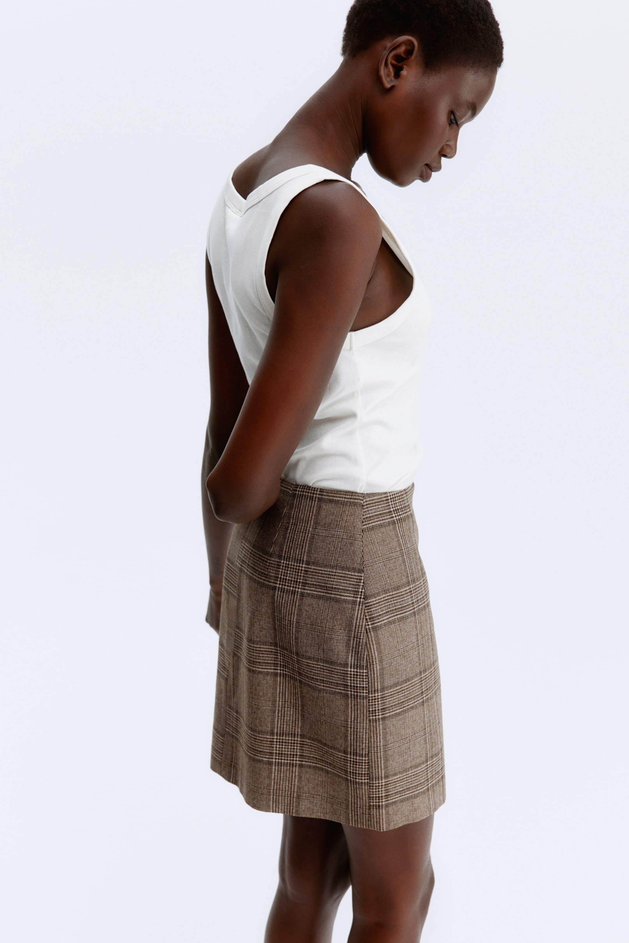 View larger image: A brown plaid mini skirt is being shown from a side and slightly back angle. The skirt is patterned with thin intersecting beige and dark brown lines, forming a check design, and is being worn with a white sleeveless top tucked in.