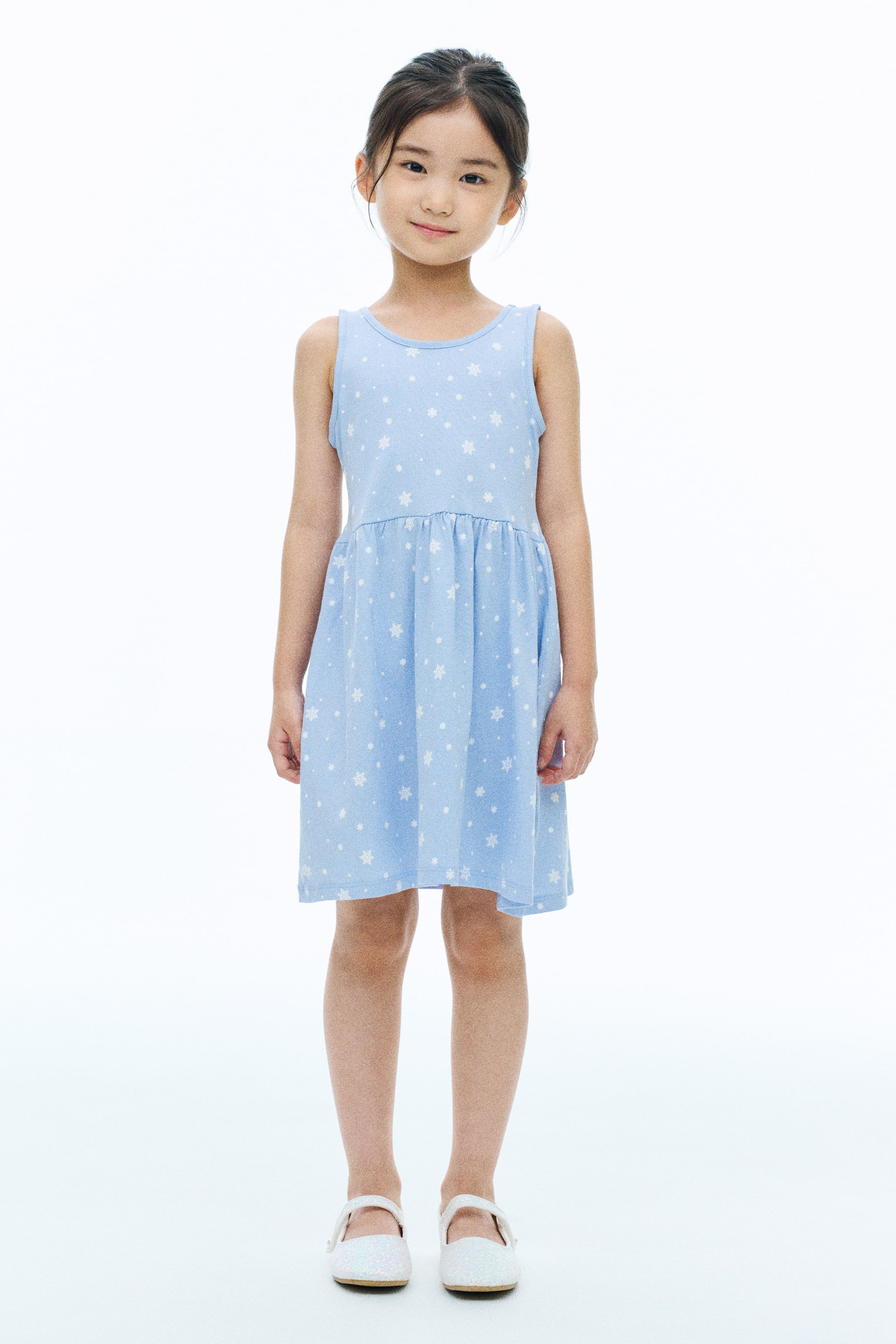 View larger image: A light blue sleeveless jersey dress featuring an all-over white snowflake print, a round neckline, a gathered waist, and a flared skirt is worn by the model. The model is standing facing forward, pairing the dress with white flat shoes.