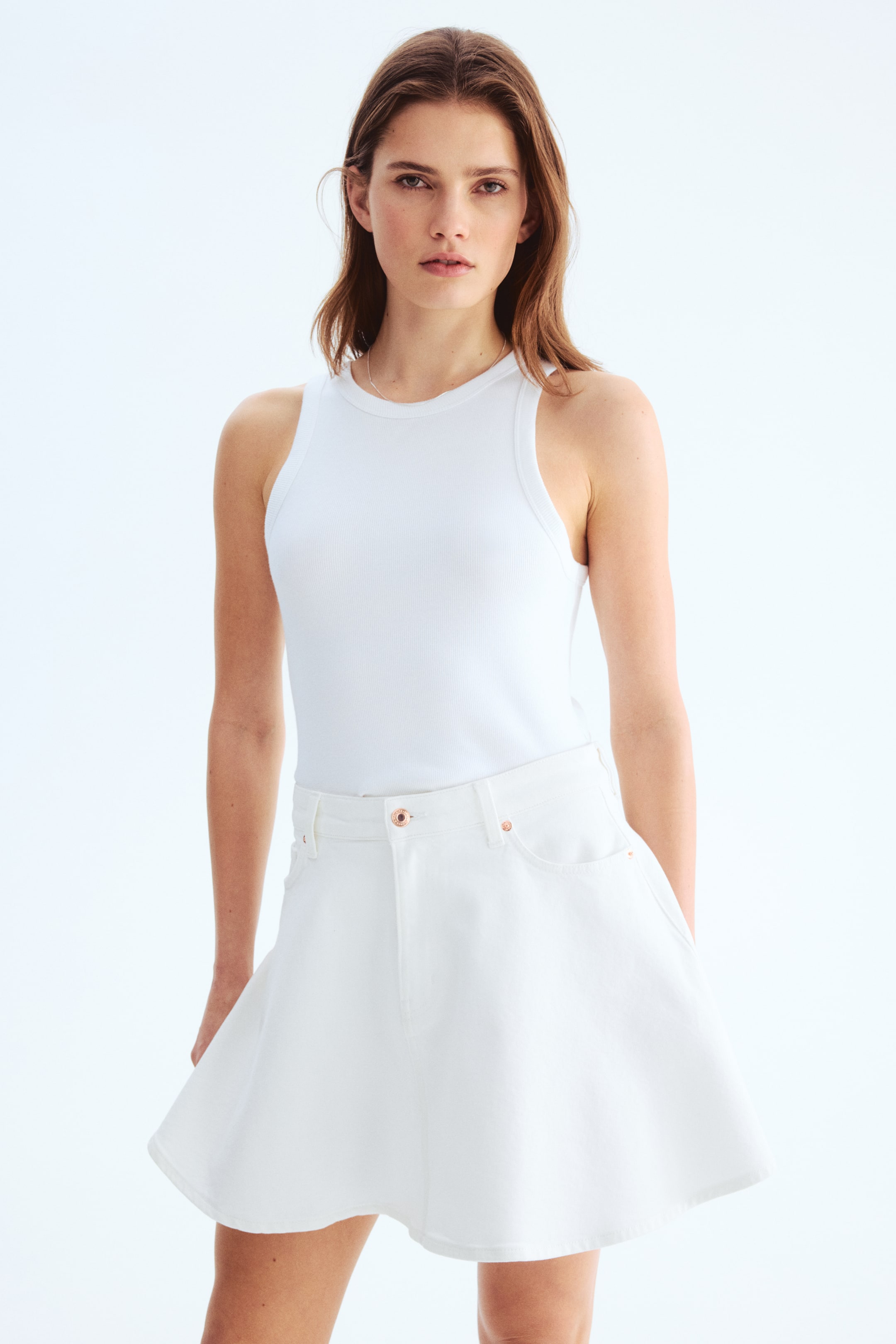 View larger image: The white ribbed tank top is showcased front-on, boasting a crew neck and broad shoulder straps, neatly tucked into a white flared mini skirt. The skirt highlights front pockets and a rose gold-toned button closure at the waist.