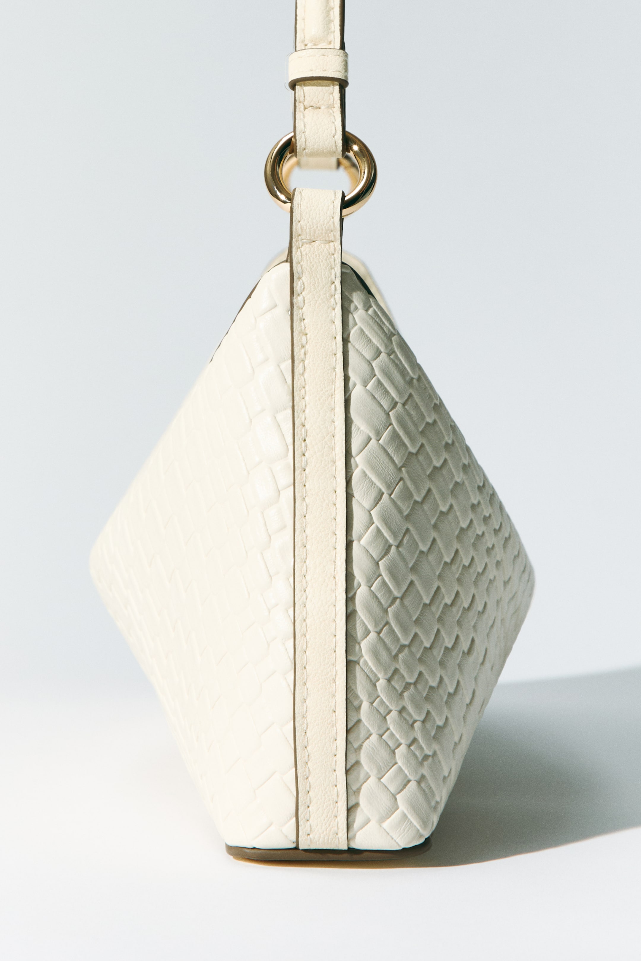 Voir une image plus grande: Un sac à main de couleur crème est suspendu verticalement, ses côtés texturés arborant un motif tissé séparé par un panneau central vertical lisse. Un anneau doré relie le sac à une fine bandoulière en haut.