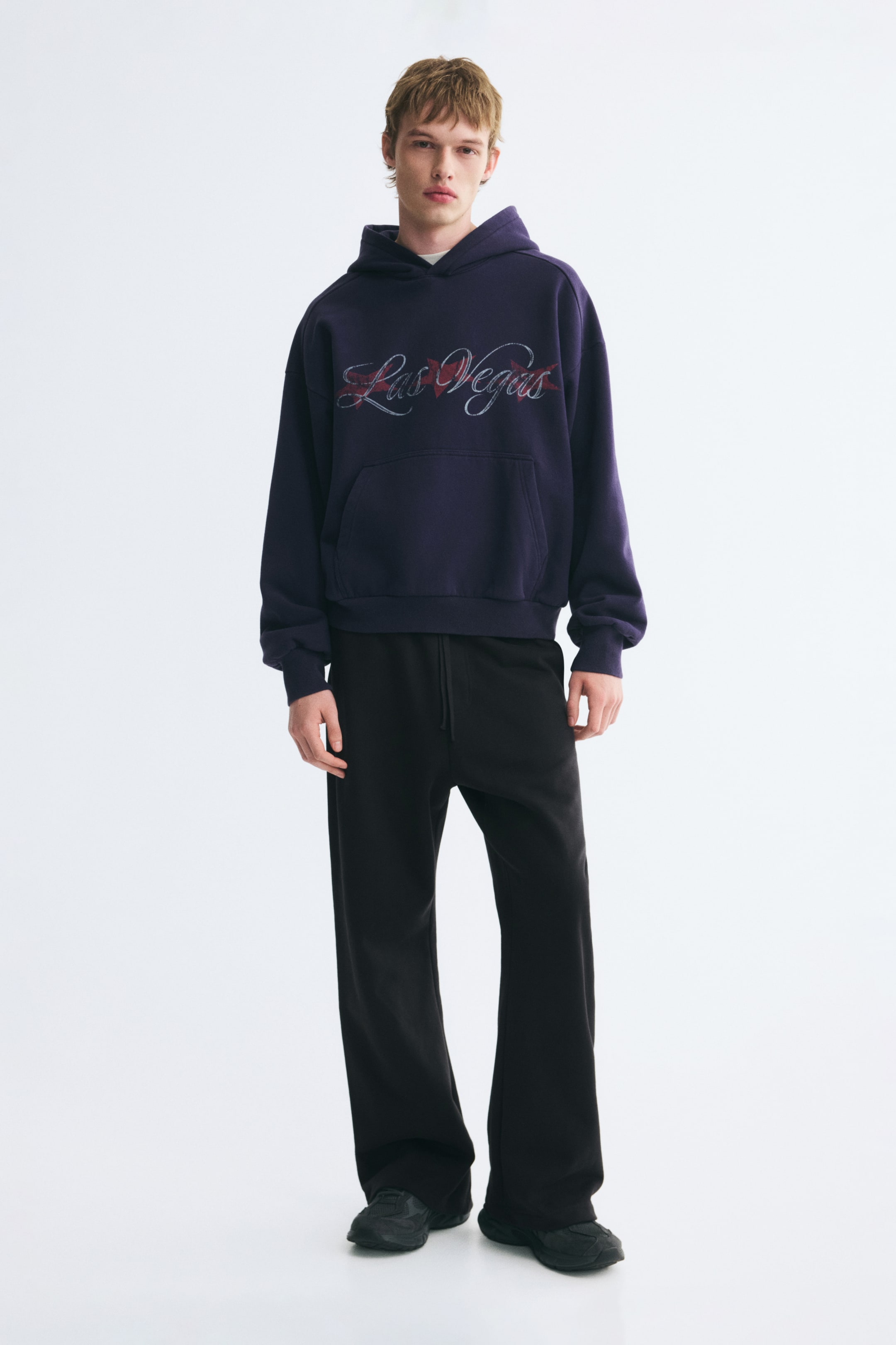 View larger image: Black track pants with a relaxed fit and straight legs are being worn by the model. The pants, which feature an elasticized drawstring waist, are styled with a dark purple graphic hoodie and black runners.