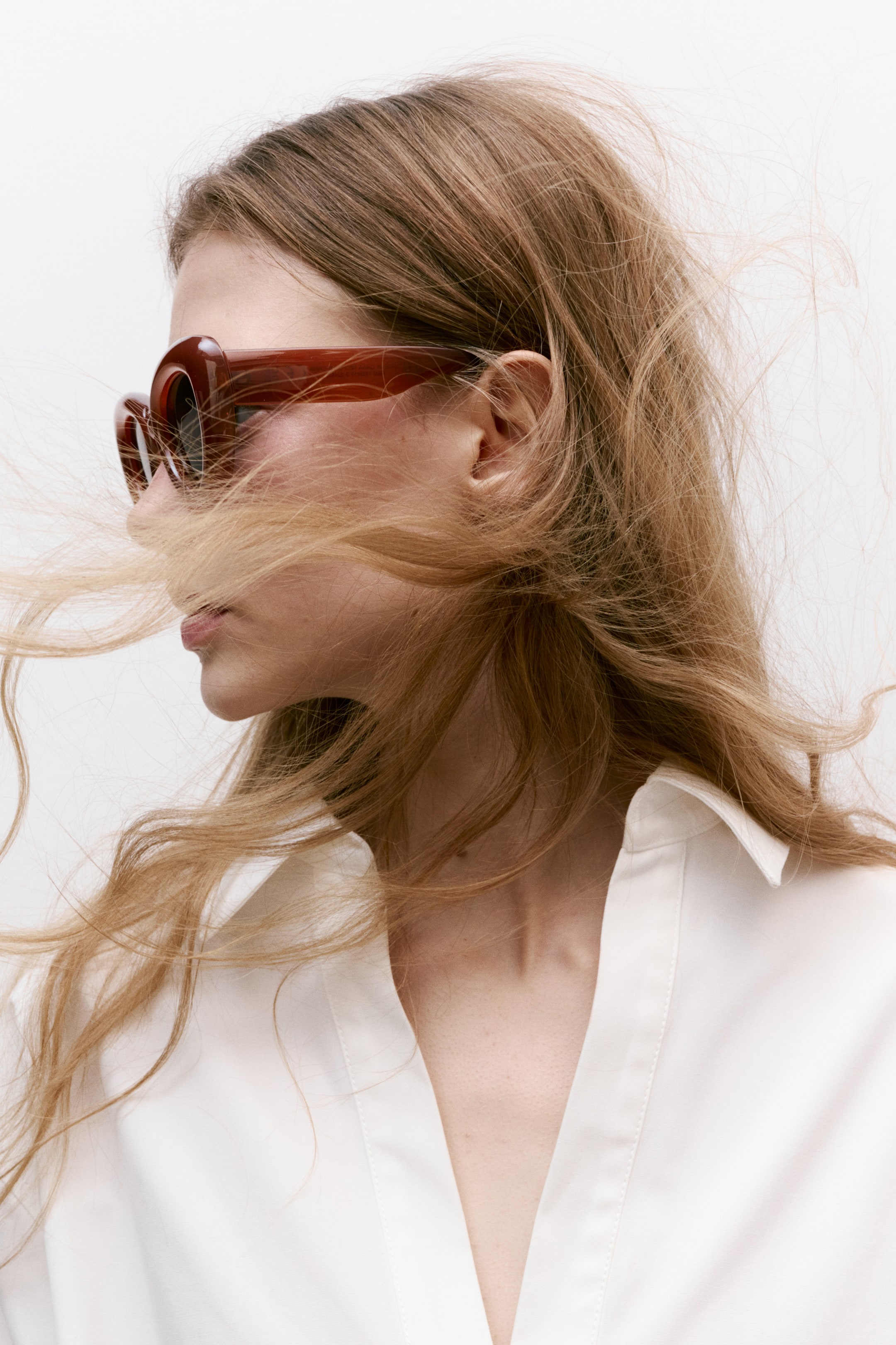 Agrandir l'image: Les lunettes de soleil surdimensionnées en écaille de tortue marron présentent des montures épaisses et incurvées, vues de côté face à gauche. Une chemise blanche à col avec un col en V pointu et ouvert est visible, révélant un tissu lisse.