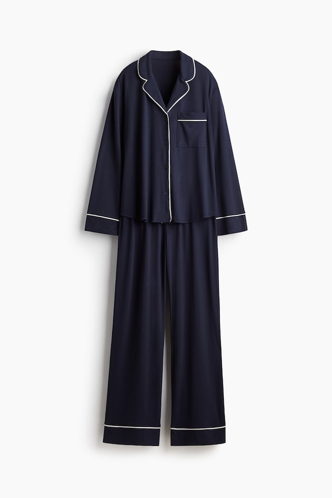 A navy blue pyjama set is presented, showing its front. The long-sleeved shirt is featuring notch lapels, white buttons down the front, and a patch pocket on the left chest. White piping is outlining the lapels, front opening, cuffs, and pocket, while the matching bottoms are featuring wide, straight legs with white piping at the hems. - 1