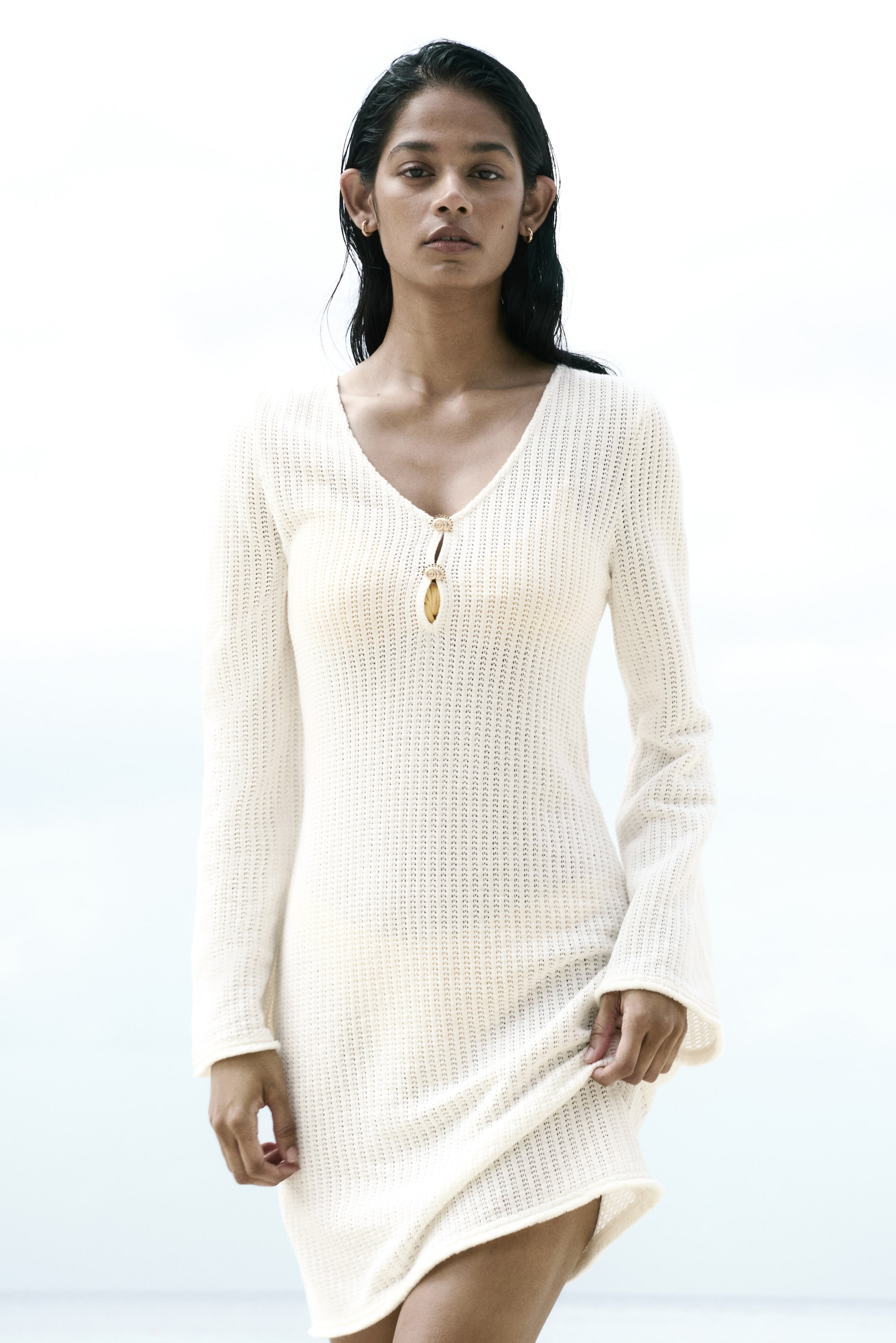 View larger image: The off-white knitted dress is displayed facing forward, showcasing a V-neckline with a keyhole detail, highlighted by two decorative gold-toned buttons. It features long, slightly flared sleeves and a body-skimming fit, with the model's hand lifting its short hem.