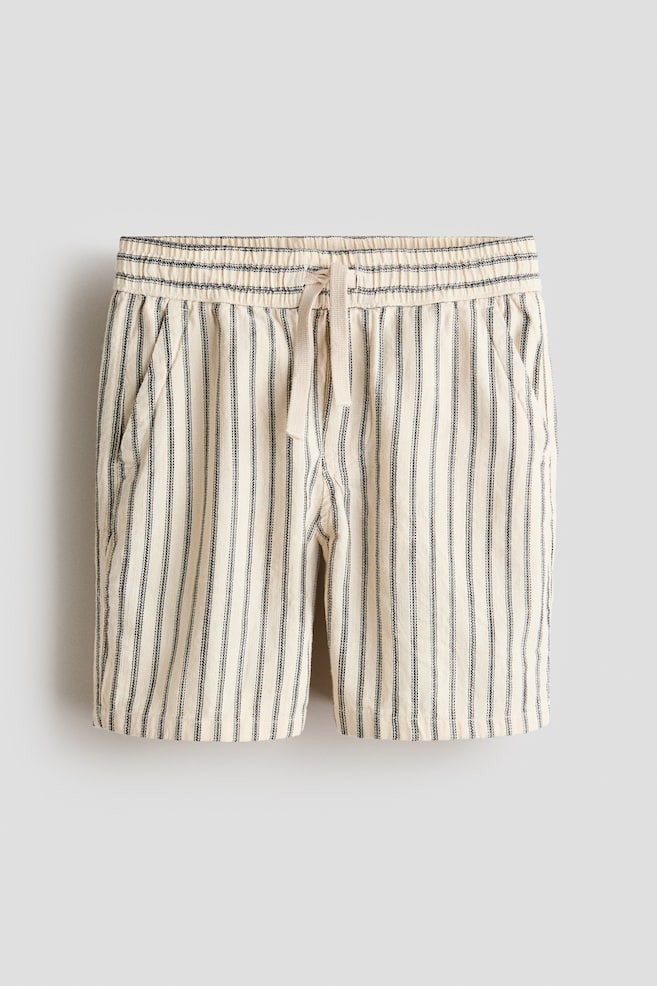 Les shorts à coupe ample présentent un tissage en coton texturé de couleur blanc cassé avec de fines rayures verticales noires. Ils sont dotés d'une taille élastiquée avec un cordon de serrage beige noué, d'une fausse braguette et de poches avant diagonales. - 1