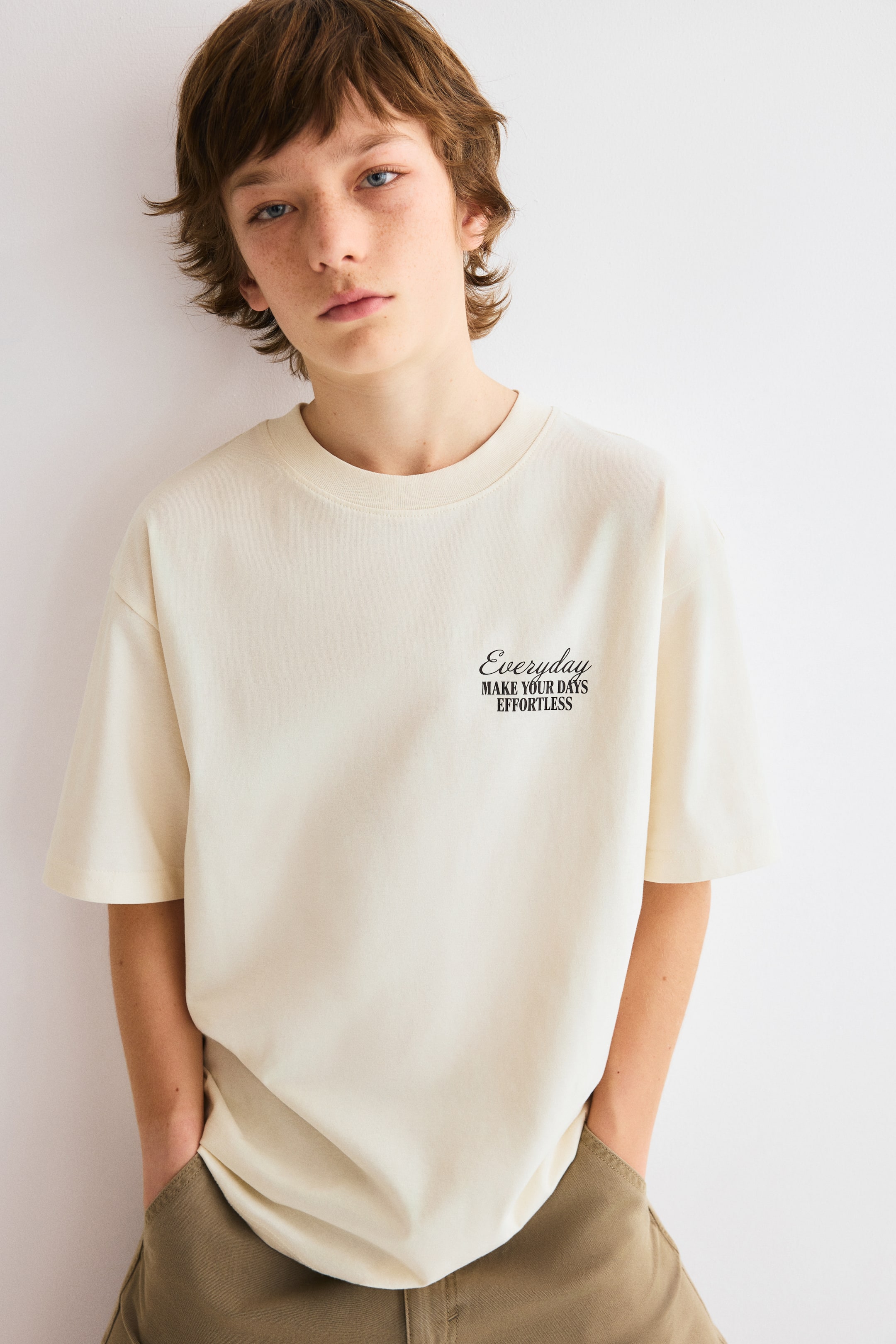 View larger image: A cream short-sleeved t-shirt, facing slightly left, showcases an oversized fit and a ribbed crew neck. Black text, "Everyday MAKE YOUR DAYS EFFORTLESS," is printed on the left chest.
