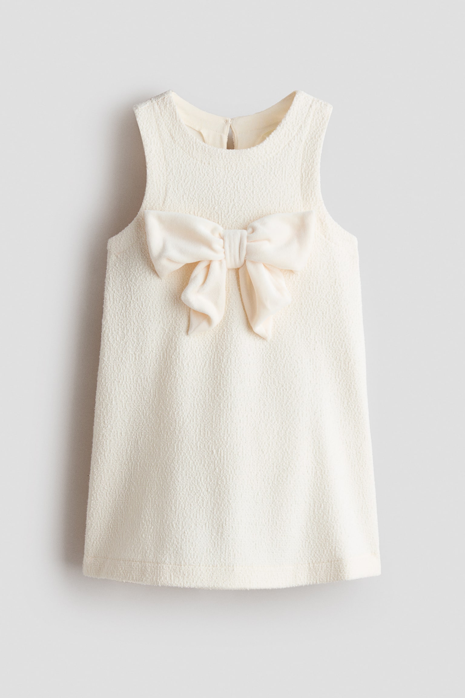 O rochie fÄrÄ mÃ¢neci, de culoare crem, este prezentatÄ din faÈÄ, avÃ¢nd un material texturat, de tip boucle, Èi un decolteu rotund. Un mare fundÄ din material, asortatÄ, de culoare alb murdar, Ã®mpodobeÈte centrul pieptului, pe silueta Ã®n A.