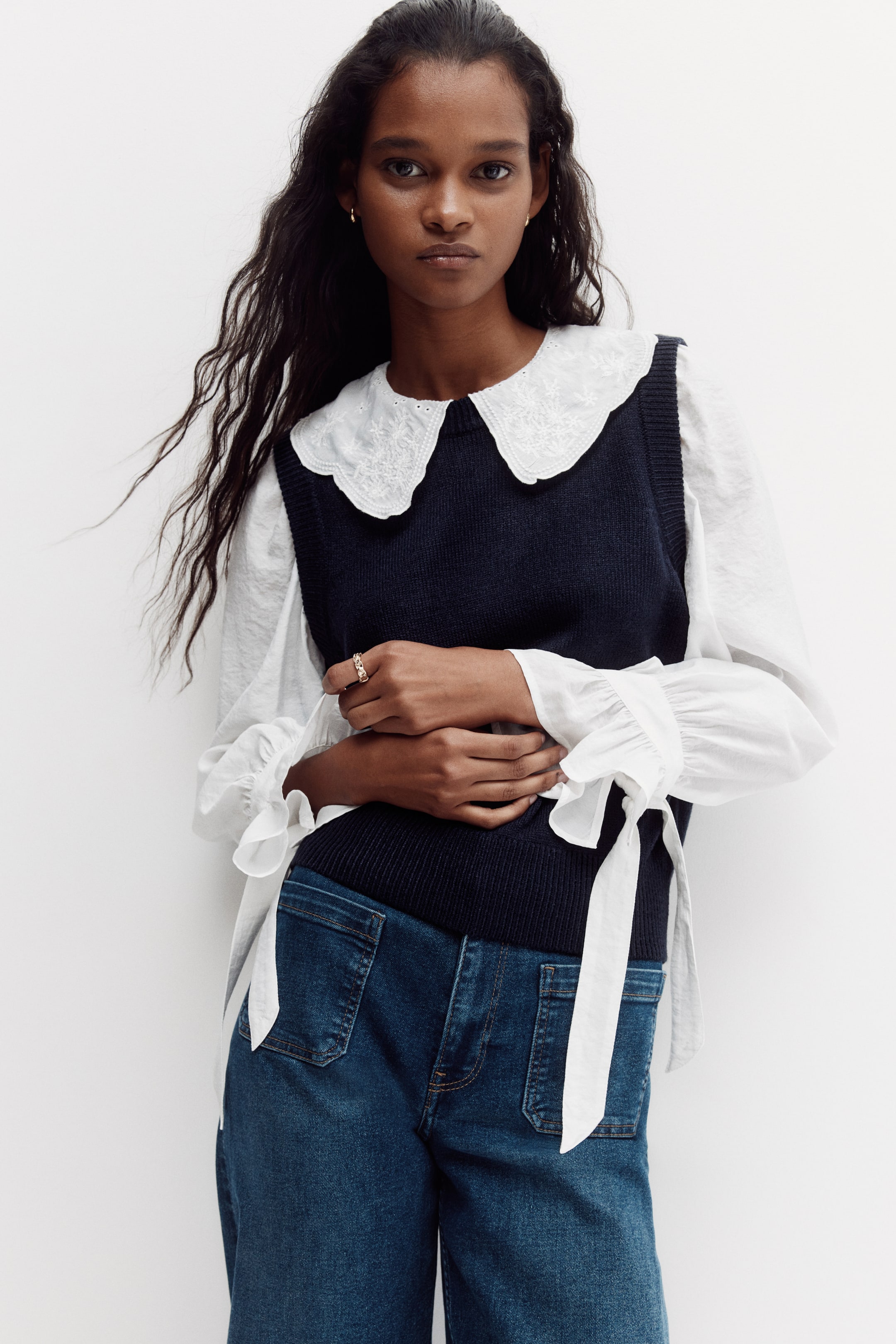 View larger image: The model wears a dark blue, fine-knit sweater vest with a round, rib-trimmed neckline, ribbed armholes and a ribbed hem. The vest is layered over a white blouse with an embroidered Peter Pan collar, and the model clasps her hands over the front.