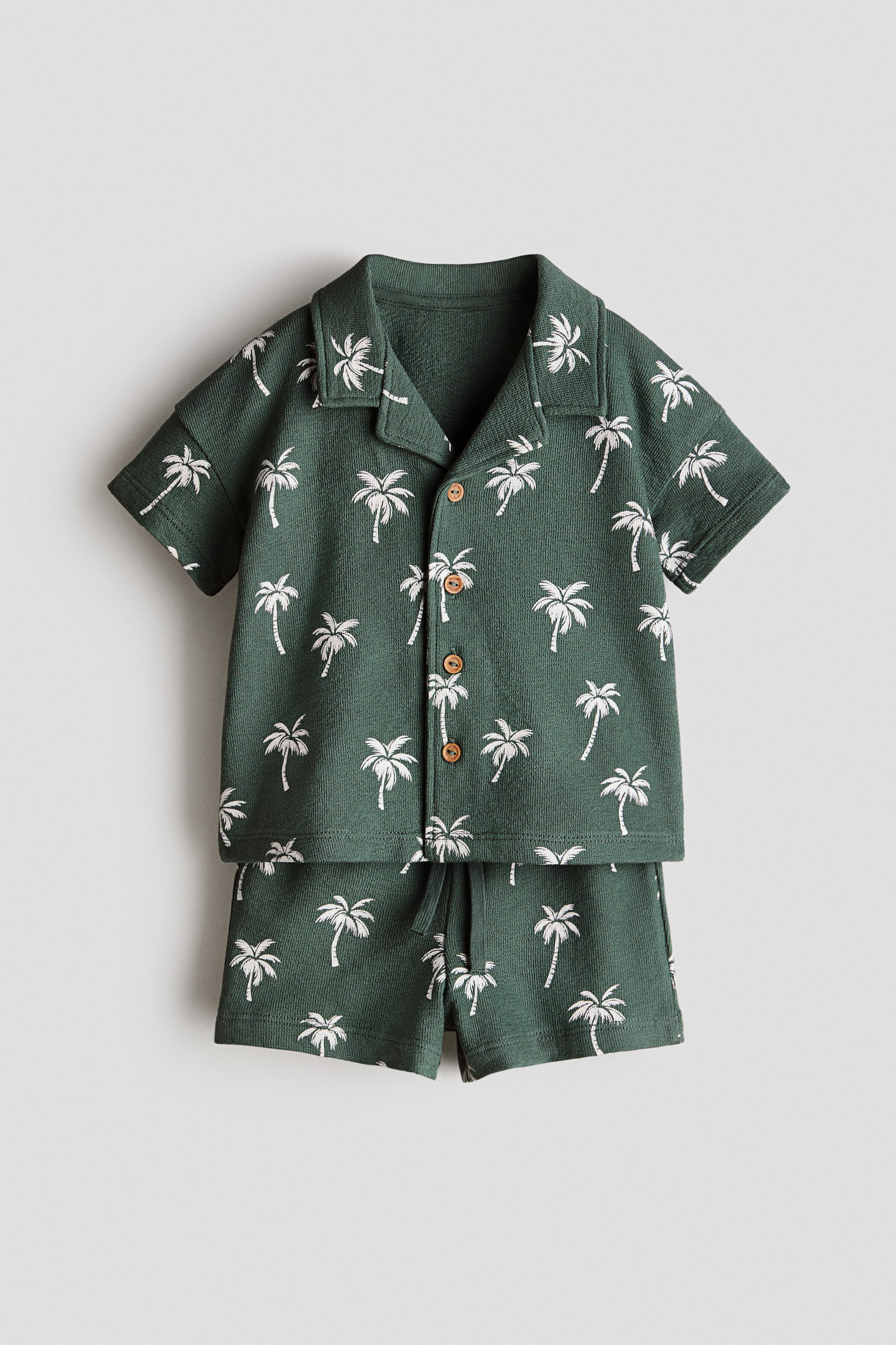 Agrandir l'image: Un ensemble vert foncé avec des motifs de palmiers blancs présente une chemise à manches courtes et un short assorti vus de face. La chemise est dotée d'un col de style resort, de trois boutons marron visibles sur le devant et d'épaules tombantes, tandis que le short affiche une taille élastiquée avec un cordon de serrage noué.
