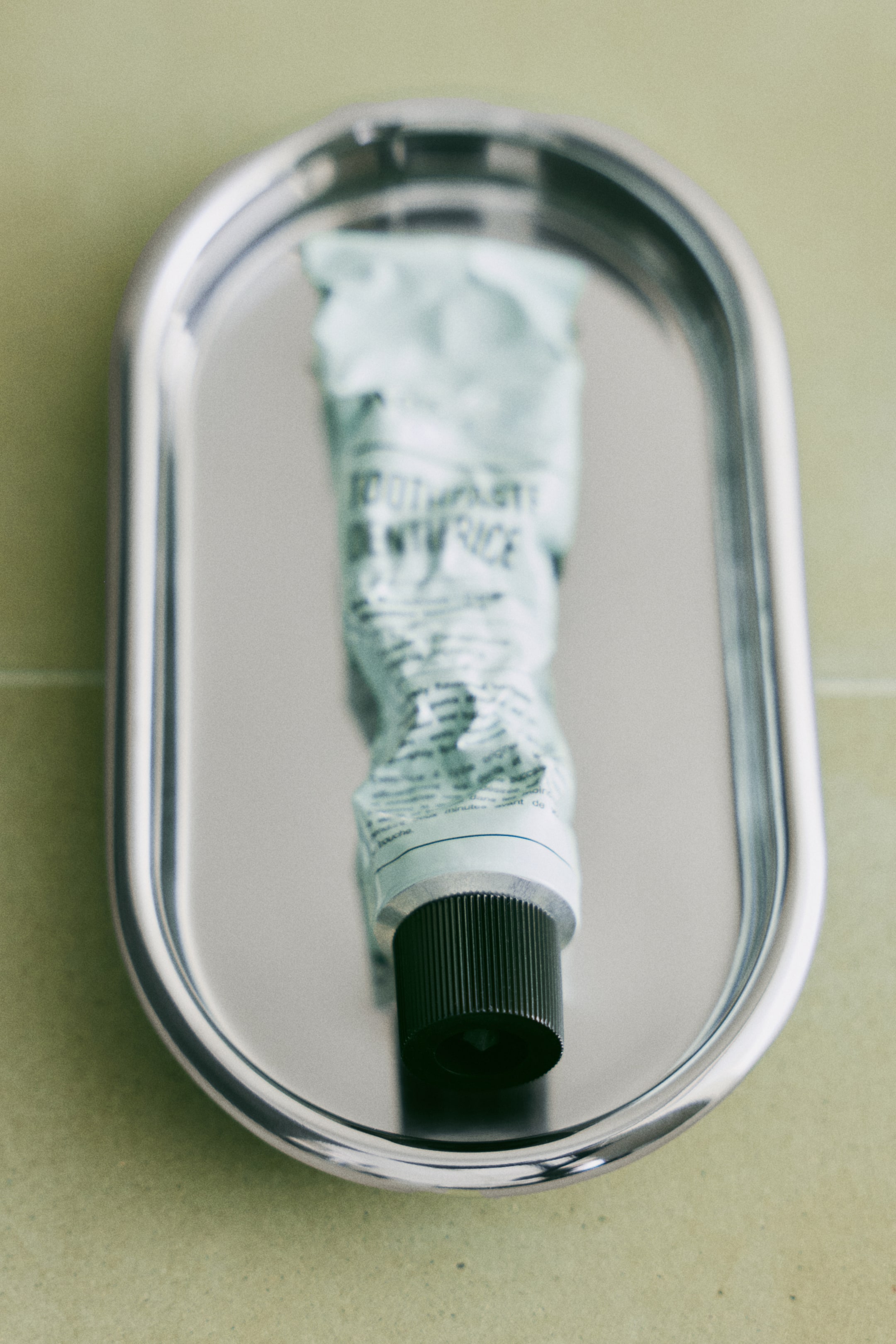 Größeres Bild ansehen: Eine hellgrüne Zahnpastatube liegt horizontal und erscheint teilweise zusammengedrückt auf einem silbernen ovalen Tablett, mit einem schwarzen gerippten Schraubverschluss und sichtbarem Text, einschließlich "toothpaste" und "dentifrice".