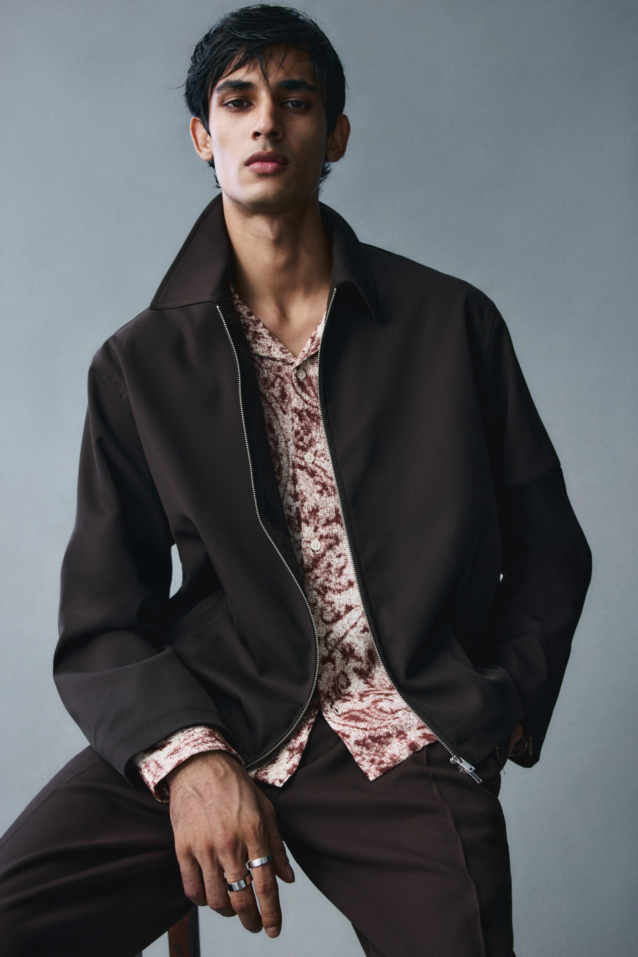 View larger image: A dark brown twill overshirt with a collar and a silver two-way zipper down the front is worn open by the model. The overshirt is layered over a patterned button-down shirt and paired with matching dark brown trousers.