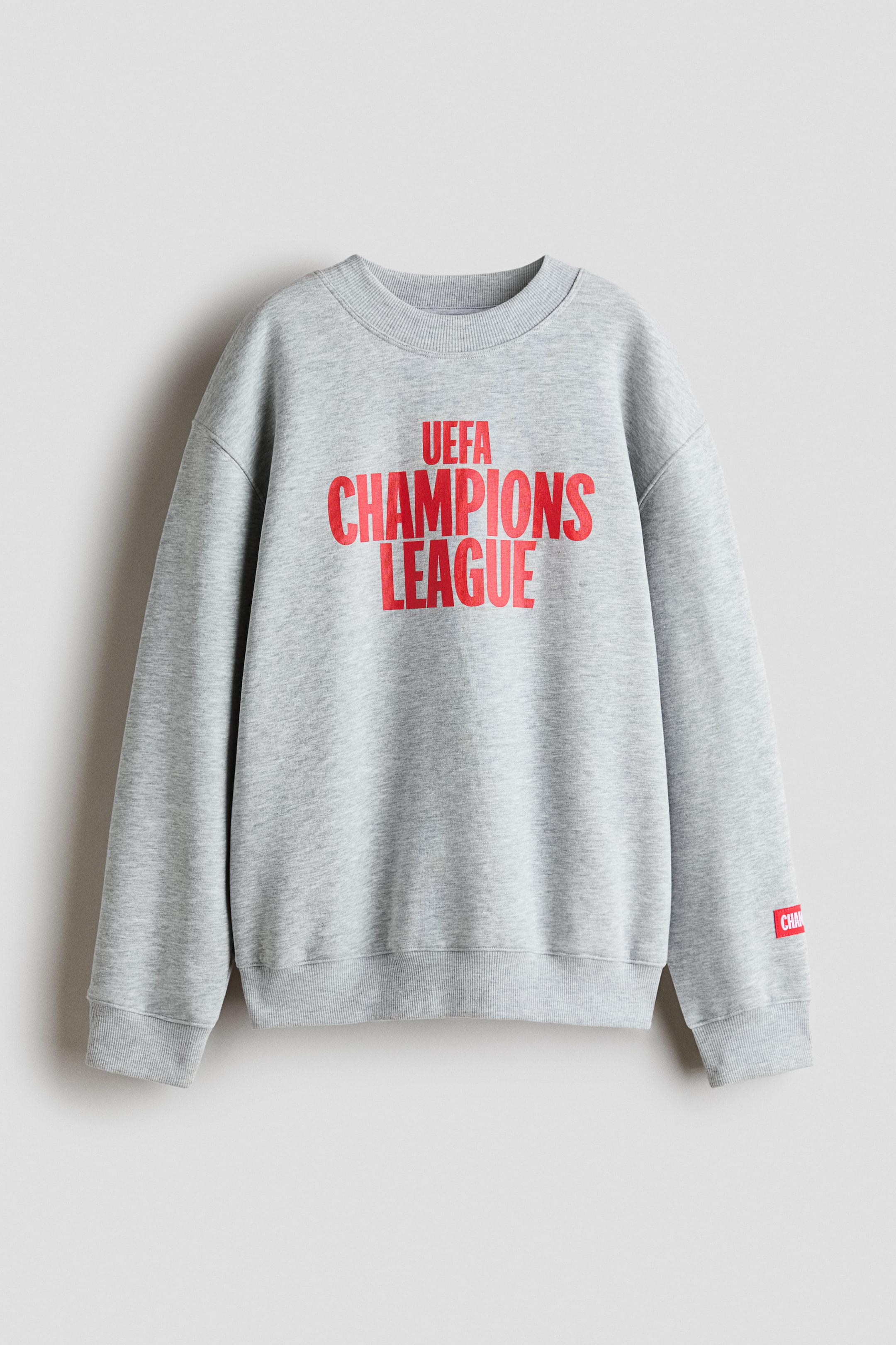Näytä suurempi kuva: Vaaleanharmaa fleece-collegepaita näkyy etupuolelta kuvattuna, ja siinä on rinnassa näyttävä punainen painatus "UEFA CHAMPIONS LEAGUE". Malli on väljä istuvuudeltaan, ja siinä on pyöreä ribbikaula-aukko, pudotetut olkapäät sekä ribbiresorit hihansuissa ja helmassa.