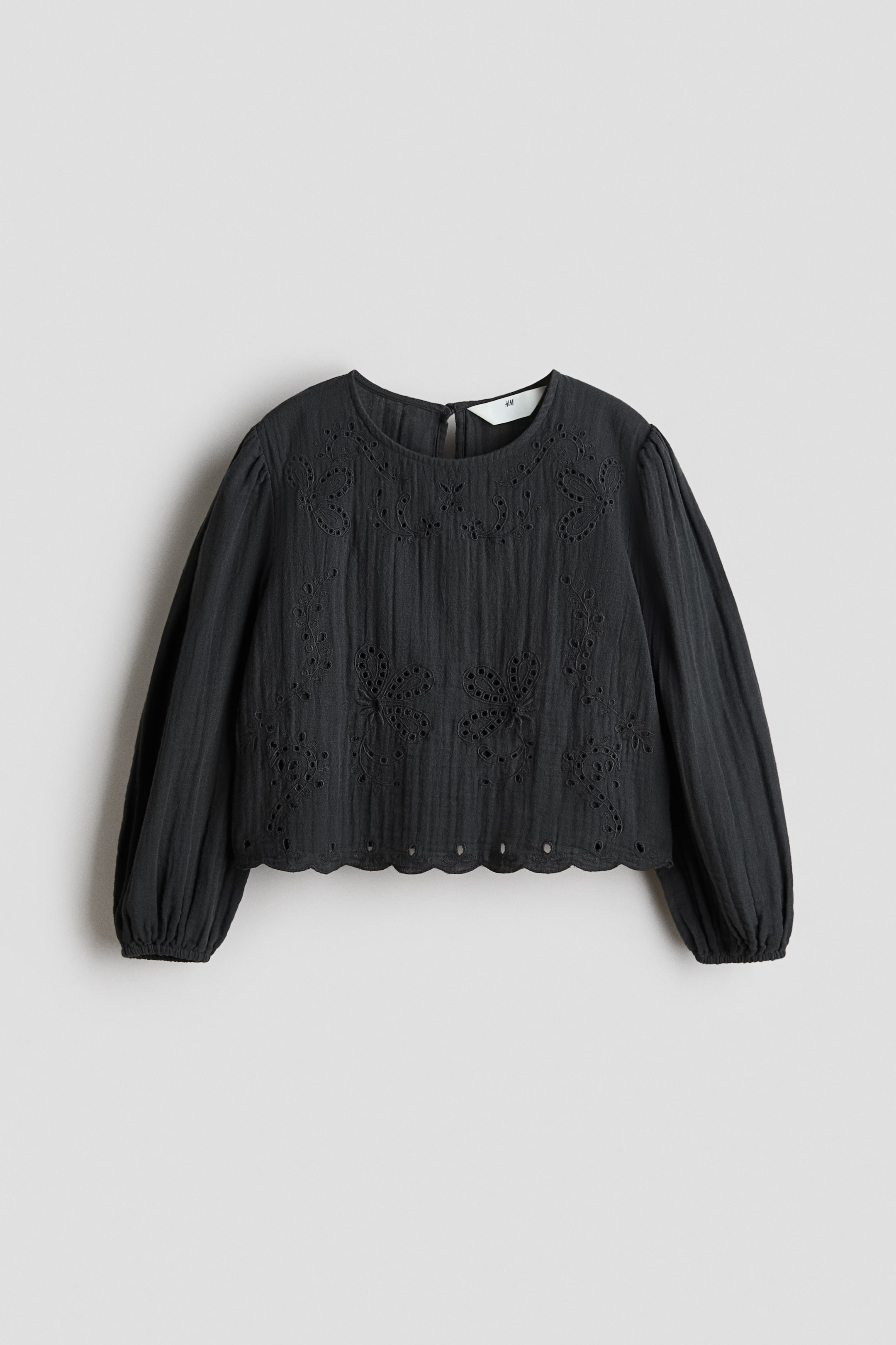 View larger image: A black cropped blouse is shown from the FRONT, featuring a round neckline and long voluminous sleeves ending in elasticated cuffs. The blouse showcases vertically textured fabric with broderie anglaise embroidery in floral patterns on the upper chest and lower body, and a scalloped hem.