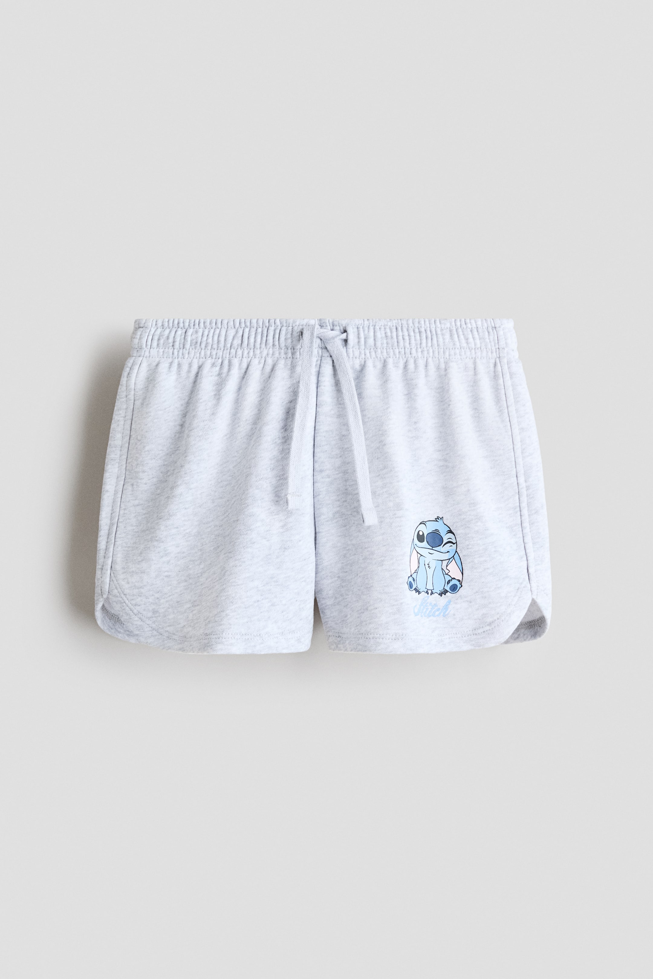 View larger image: Light grey relaxed-fit shorts in sweatshirt fabric are showcasing a frontal view, featuring an elasticated drawstring waist and a blue character print motif on the lower left leg. The shorts are crafted with gently curved hems.