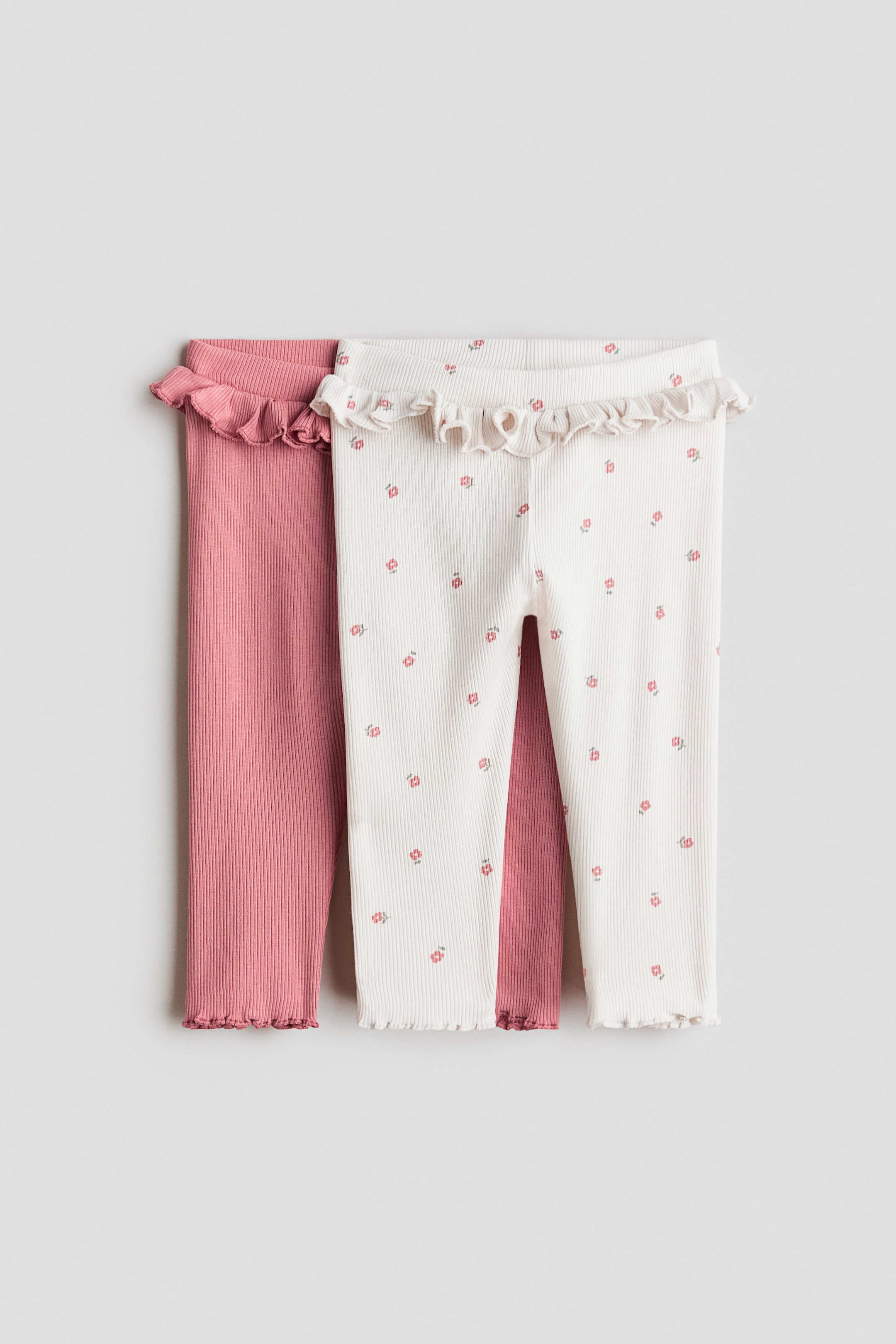 Größeres Bild ansehen: Zwei Paar gerippte Baby-Leggings werden flach liegend von vorne präsentiert. Ein Paar ist cremeweiß mit einem kleinen rosa Blumenmuster und verfügt über einen gerafften Bund sowie Lettuce-Edge-Säume. Das zweite Paar ist unifarben staubrosa und verfügt ebenfalls über einen gerafften Bund sowie Lettuce-Edge-Säume.