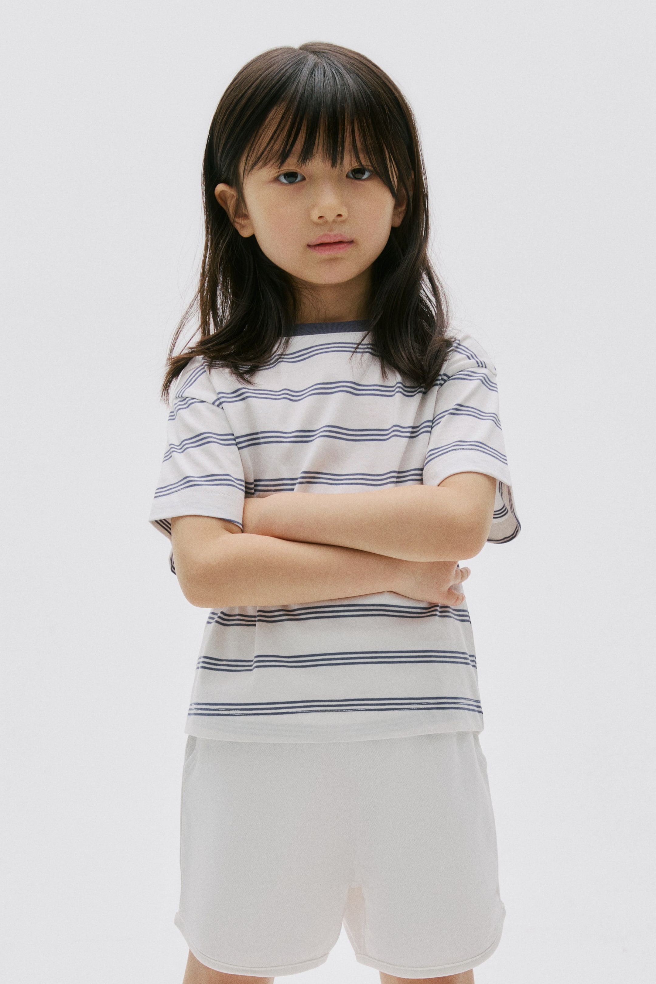 View larger image: A white short-sleeved t-shirt, featuring thin blue horizontal stripes and a blue crew neck, is being worn by a child facing forward. The t-shirt is showcasing a relaxed fit and is being paired with white mid-thigh shorts that are appearing loose.