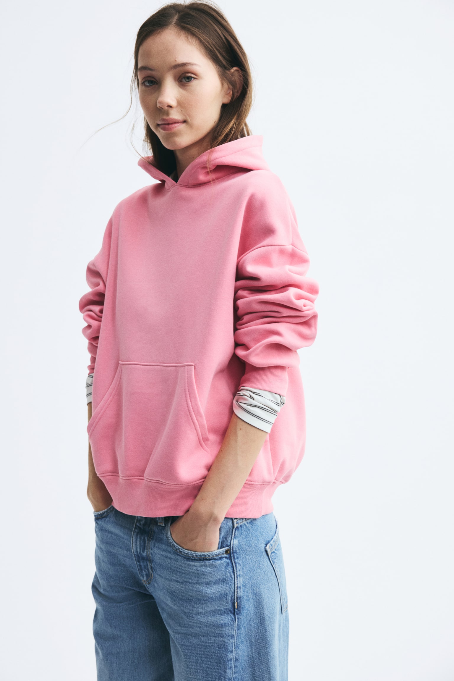 A pink fleece hoodie featuring dropped shoulders, a kangaroo pocket, and a ribbed hem is being worn by the model over a striped long-sleeve shirt. The model is standing with hands placed in the pockets of blue jeans. - 1