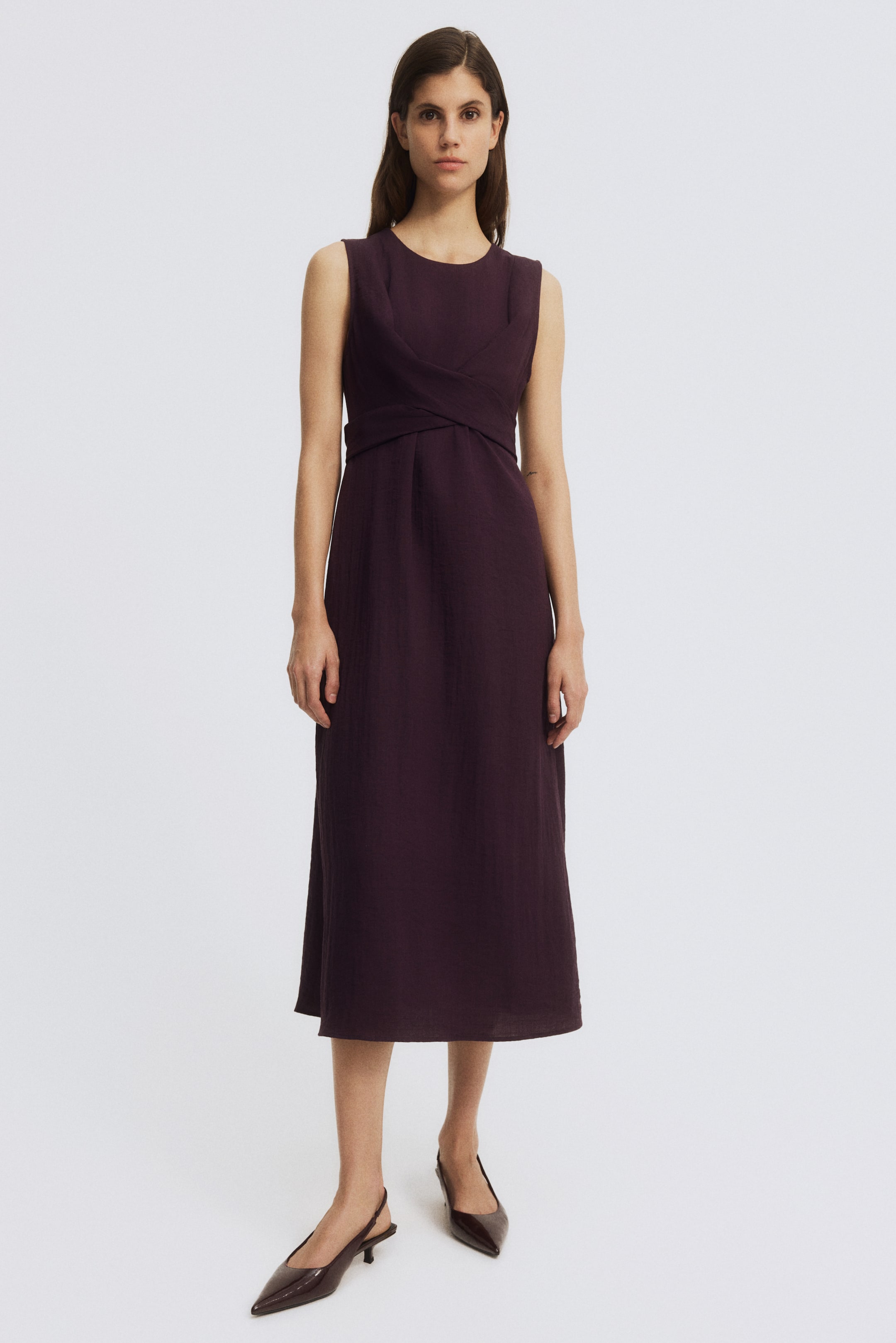View larger image: The dark plum sleeveless dress showcases a round neckline and a bodice with criss-cross wrapping, flowing into a midi-length A-line skirt.