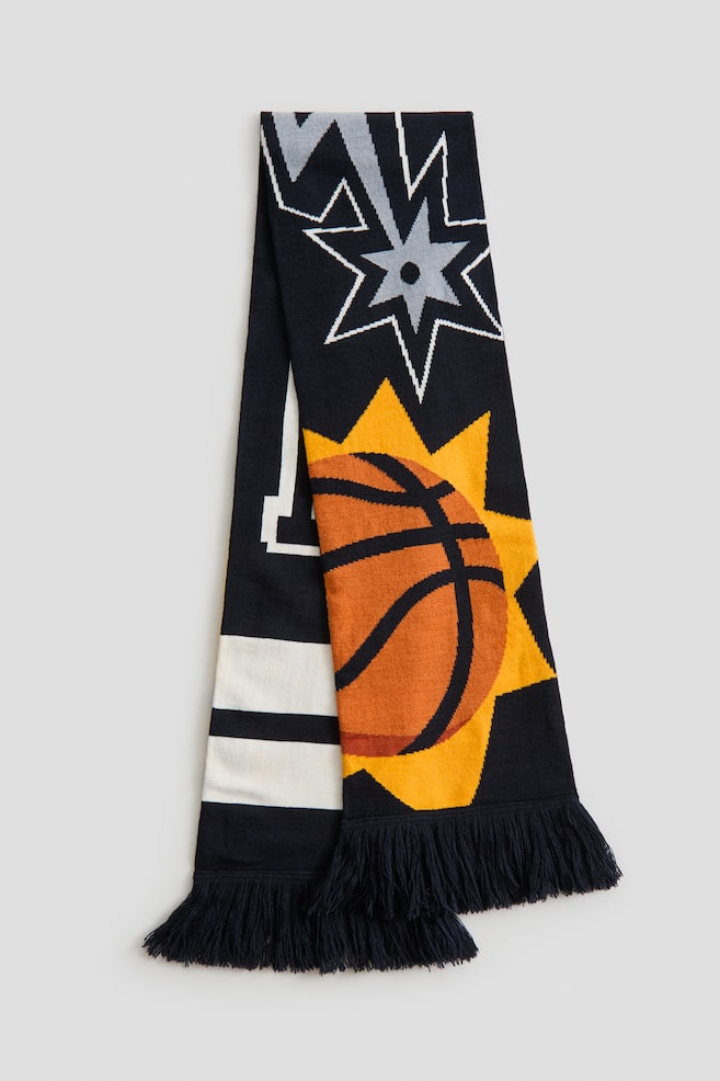 A black, white, gray, orange, and yellow knitted scarf is lying flat, folded lengthwise, presenting its front. The scarf is featuring a stylized gray star design with white outlines and white zigzag lines at its upper section, and a large orange basketball on a yellow starburst shape at its lower section. Black fringe is finishing both ends, and white horizontal stripes are visible along the folded left edge. - 1