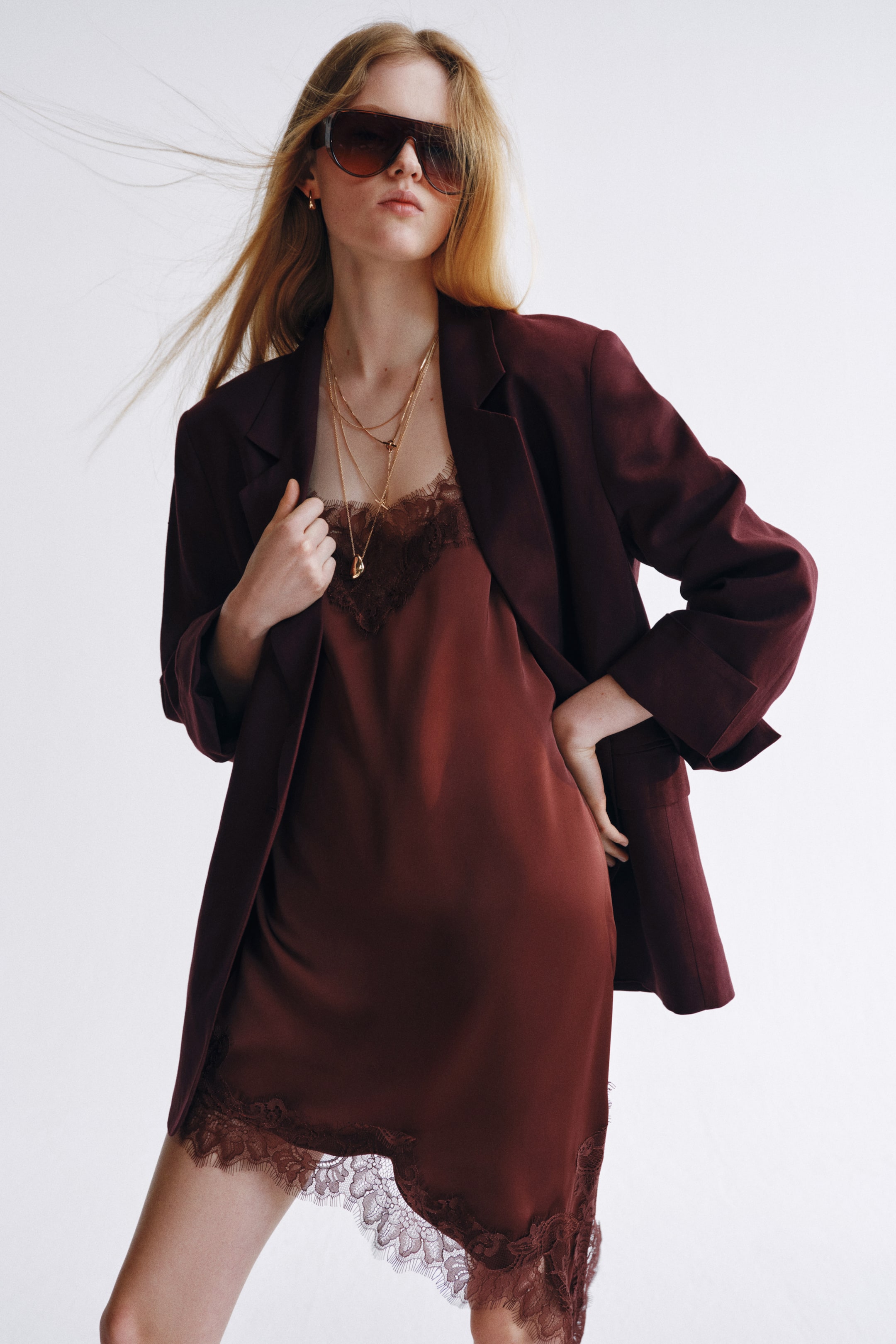 View larger image: The model is wearing an open dark red single-breasted blazer with notch lapels and long sleeves. She holds the left lapel with one hand, revealing a dark red slip dress with lace trim underneath.