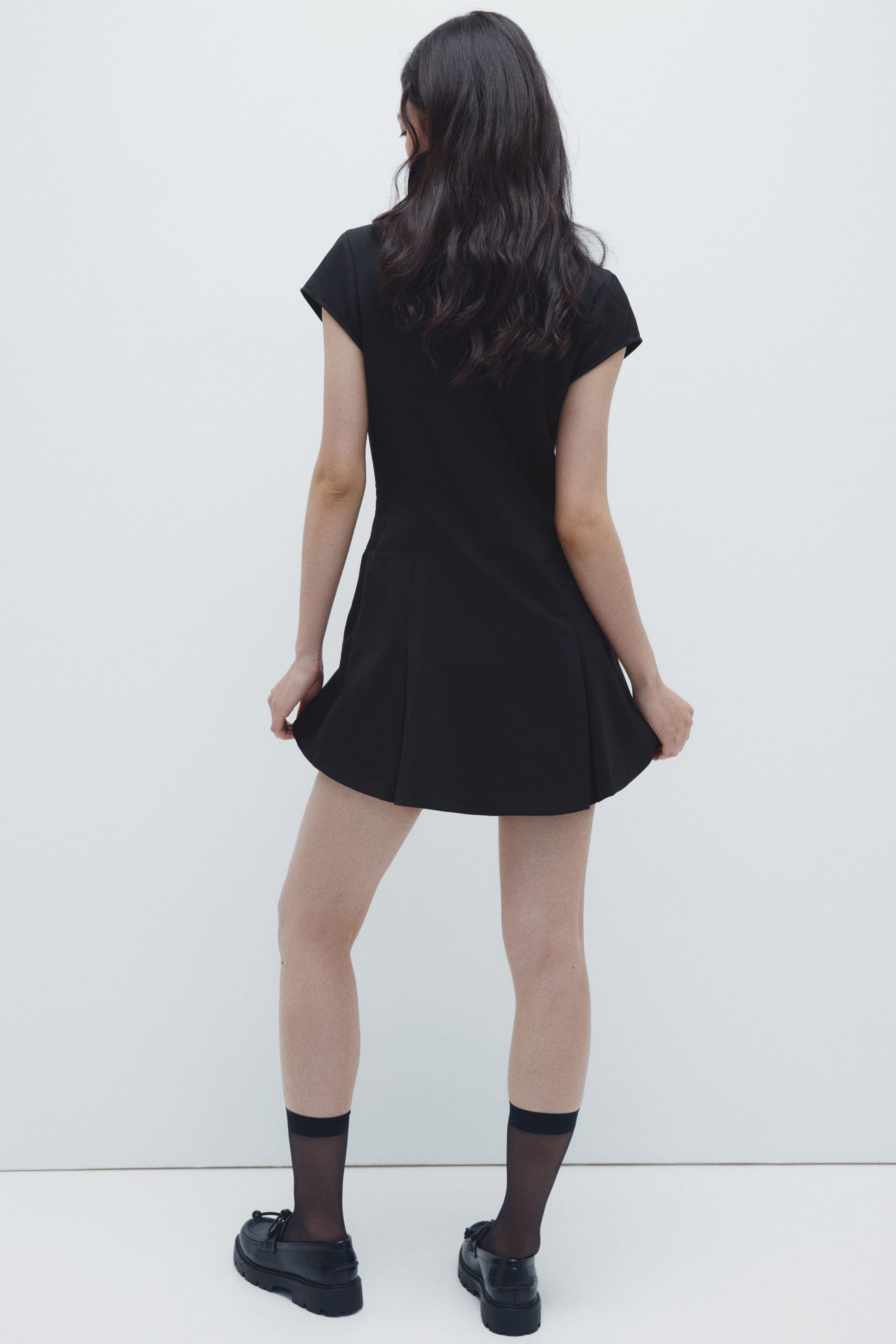 拡大画像を表示: 黒いドレスは後ろから見せられており、キャップスリーブとぴったりとしたボディスを備えています。短いプリーツスカートは広がっており、手がそっと裾を持ち上げてその動きを強調しています。