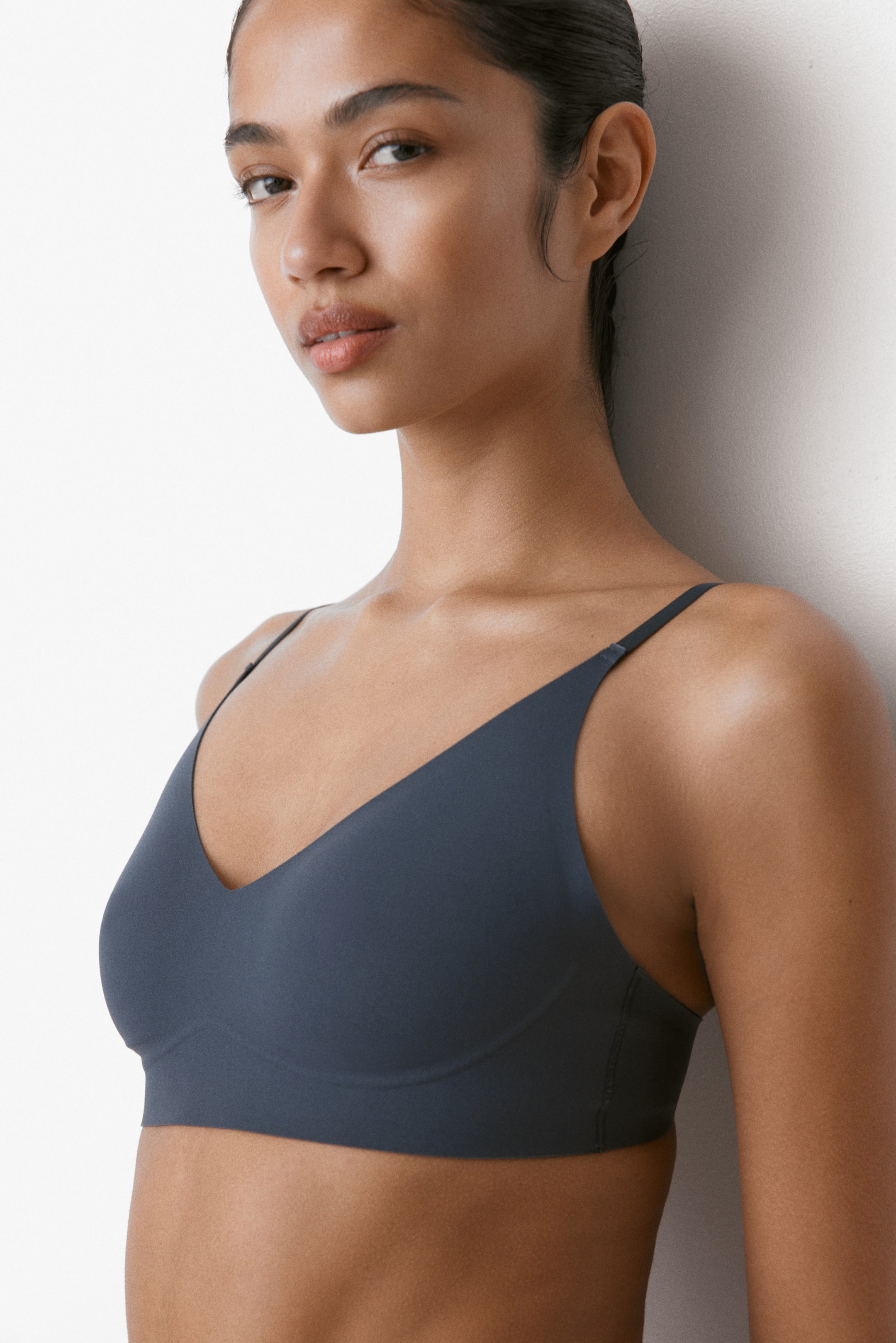 View larger image: A dark grey soft bra is shown being worn, presenting a smooth, seamless appearance with a deep V-neckline and thin spaghetti straps. It features soft cups and a wide underband, shown from the front-left angle.