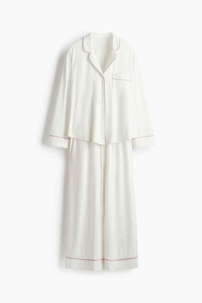 A cream-colored pyjama set is presented from the FRONT. The long-sleeved shirt is featuring notch lapels, a full button-down front, and a single chest pocket, all detailed with thin light brown piping. The matching bottoms are displaying wide, straight legs with coordinating light brown piping along the hems. - 1