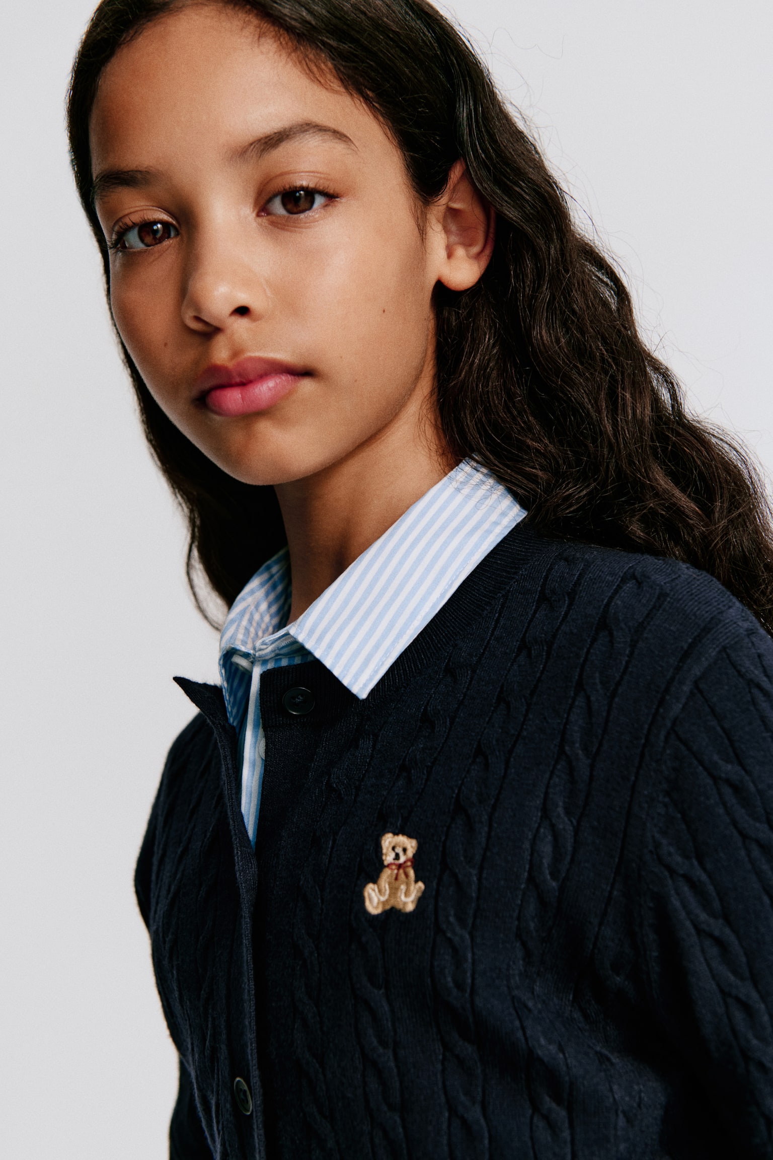 A dark blue cable knit cardigan is laid flat, front-facing, featuring a round, rib-trimmed neckline and six buttons down the front. A small embroidered teddy bear detail appears on the left chest, while long sleeves with ribbed cuffs and a ribbed hem complete the design. - 1