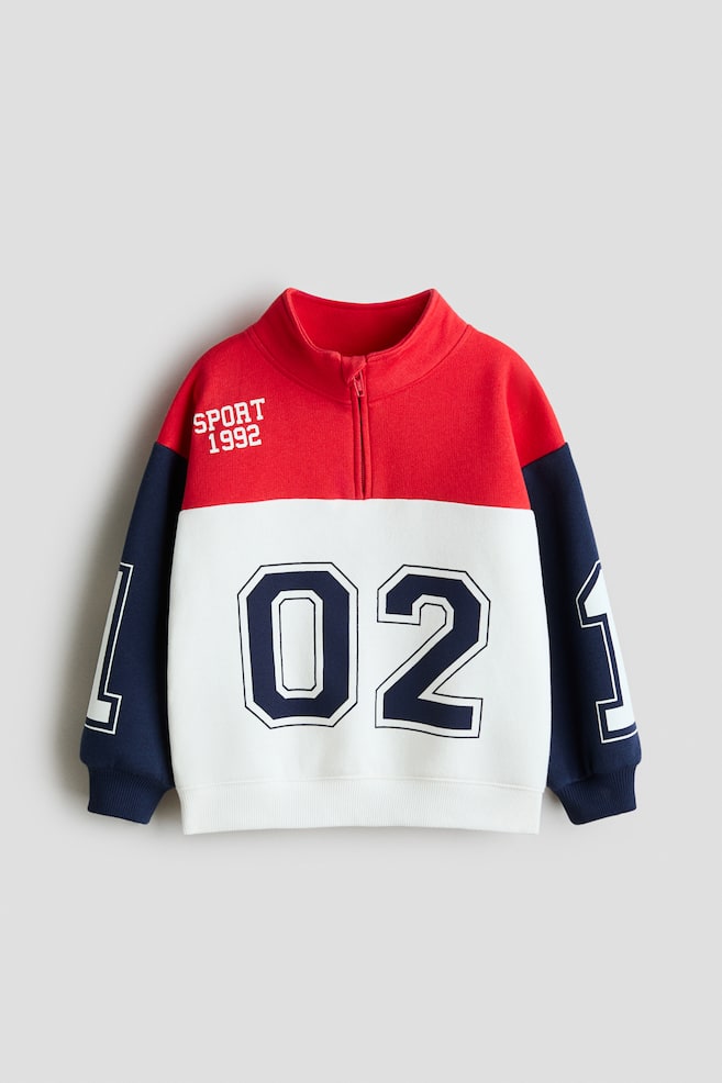 A front-facing colour-block sweatshirt features a red upper section, a white mid-section with a large navy "02" graphic, and navy sleeves with partial "1" graphics. It has a quarter-zip stand-up collar, "SPORT 1992" text on the left chest, and ribbed cuffs and hem. - 1