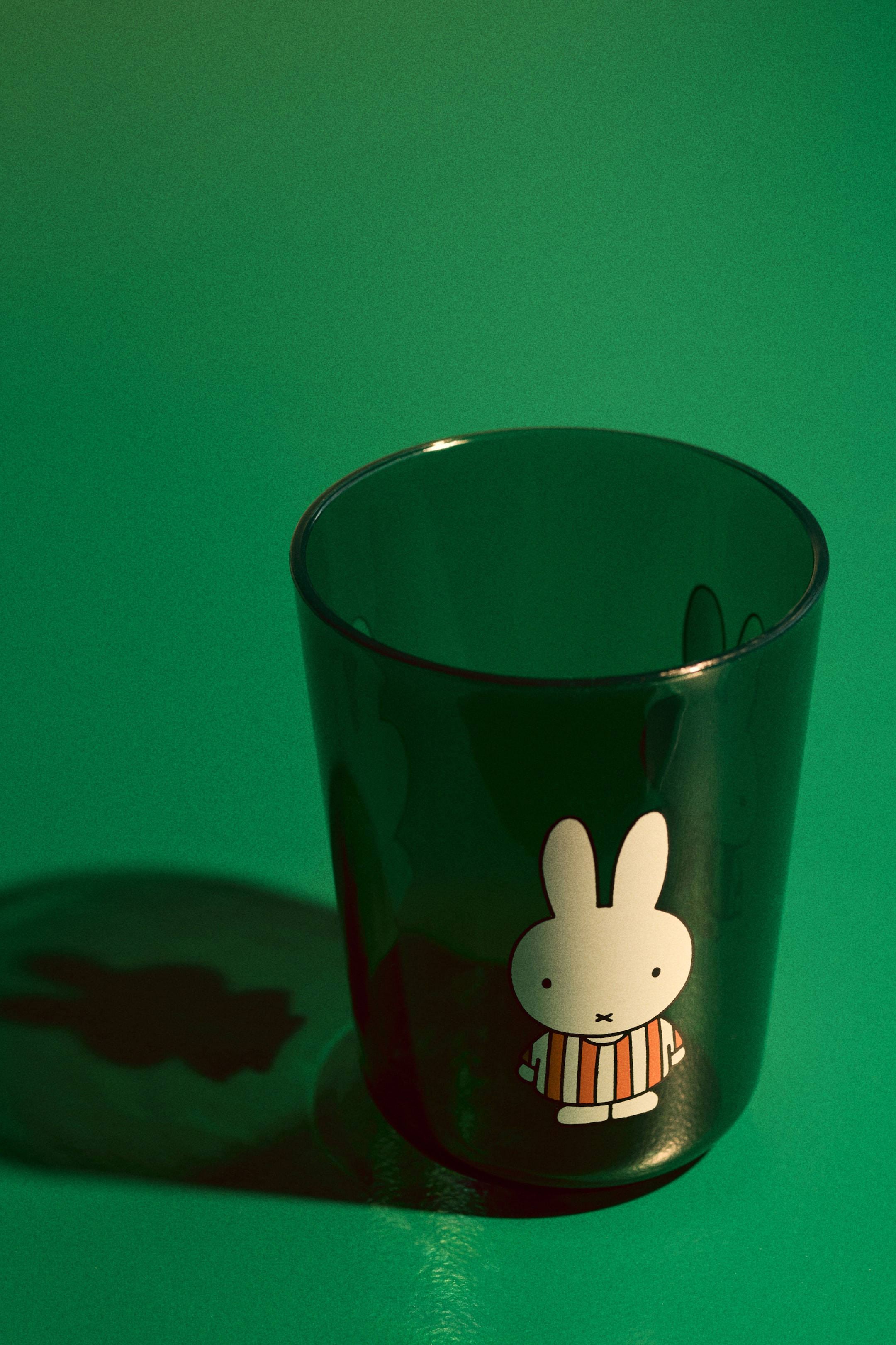 View larger image: A dark green translucent cup features a white Miffy character with black eyes and mouth, dressed in an orange and white striped shirt on its front.