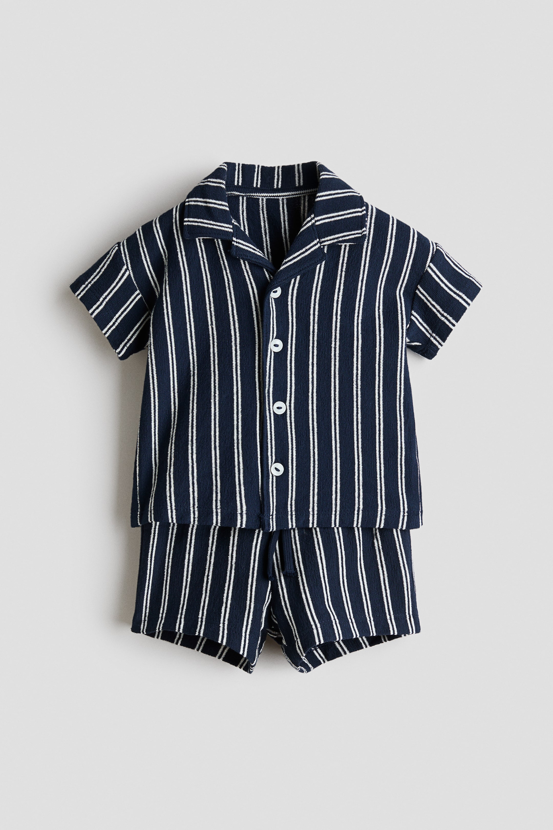 Ver imagen más grande: Un conjunto de camisa y pantalones cortos a rayas en azul marino y blanco, confeccionado en una tela ligeramente texturizada, se presenta de frente. La camisa de manga corta presenta un cuello cubano, botones blancos en la parte delantera y hombros caídos. Los pantalones cortos a juego muestran una cintura elástica.