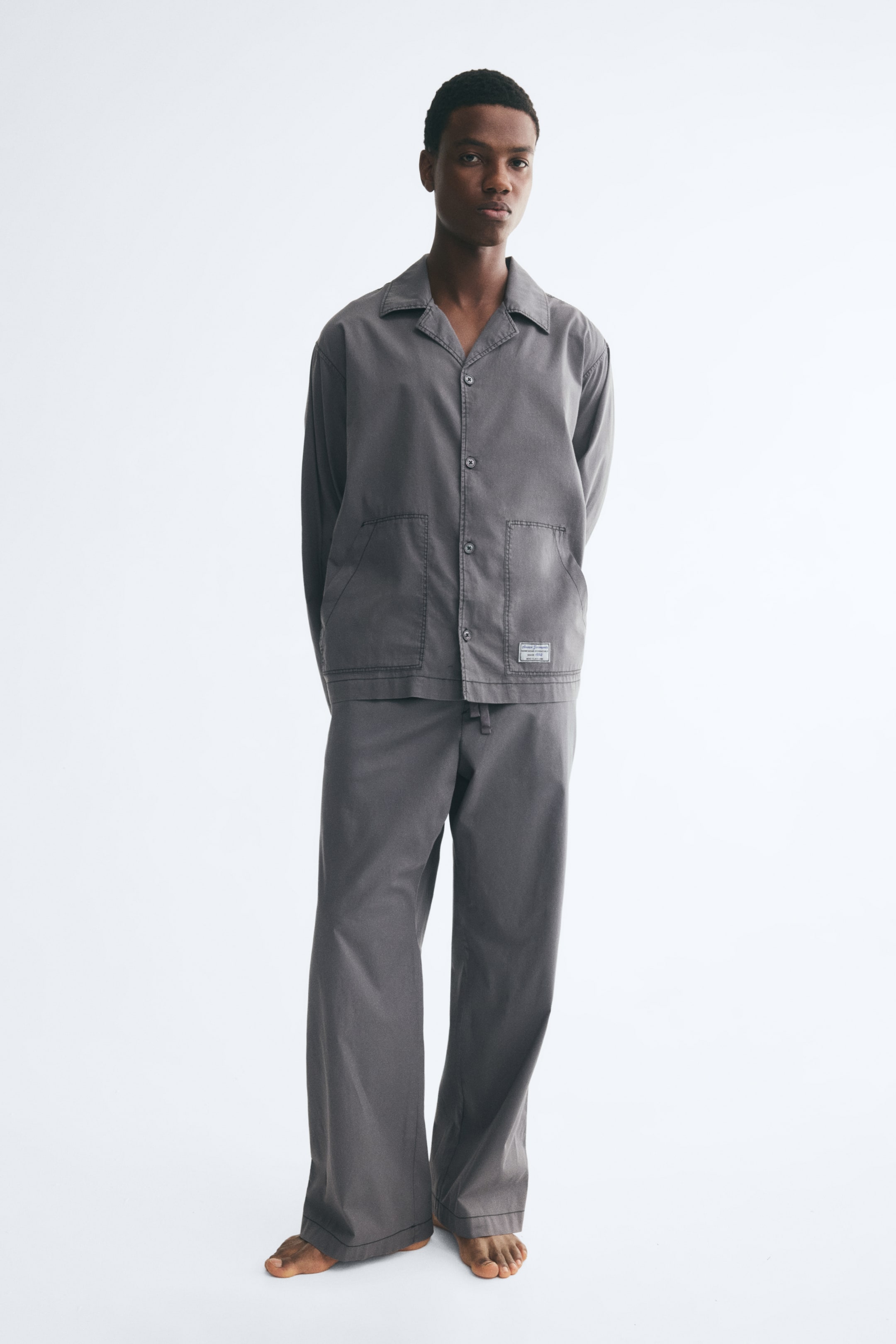 Ver imagem maior: A modelo está a usar um conjunto de pijama em popelina de algodão cinzento, com uma camisa de manga comprida, gola resort, fecho frontal por botões e bolsos de chapa à frente. As calças a condizer, de perna direita, apresentam um cordão ajustável na cintura.